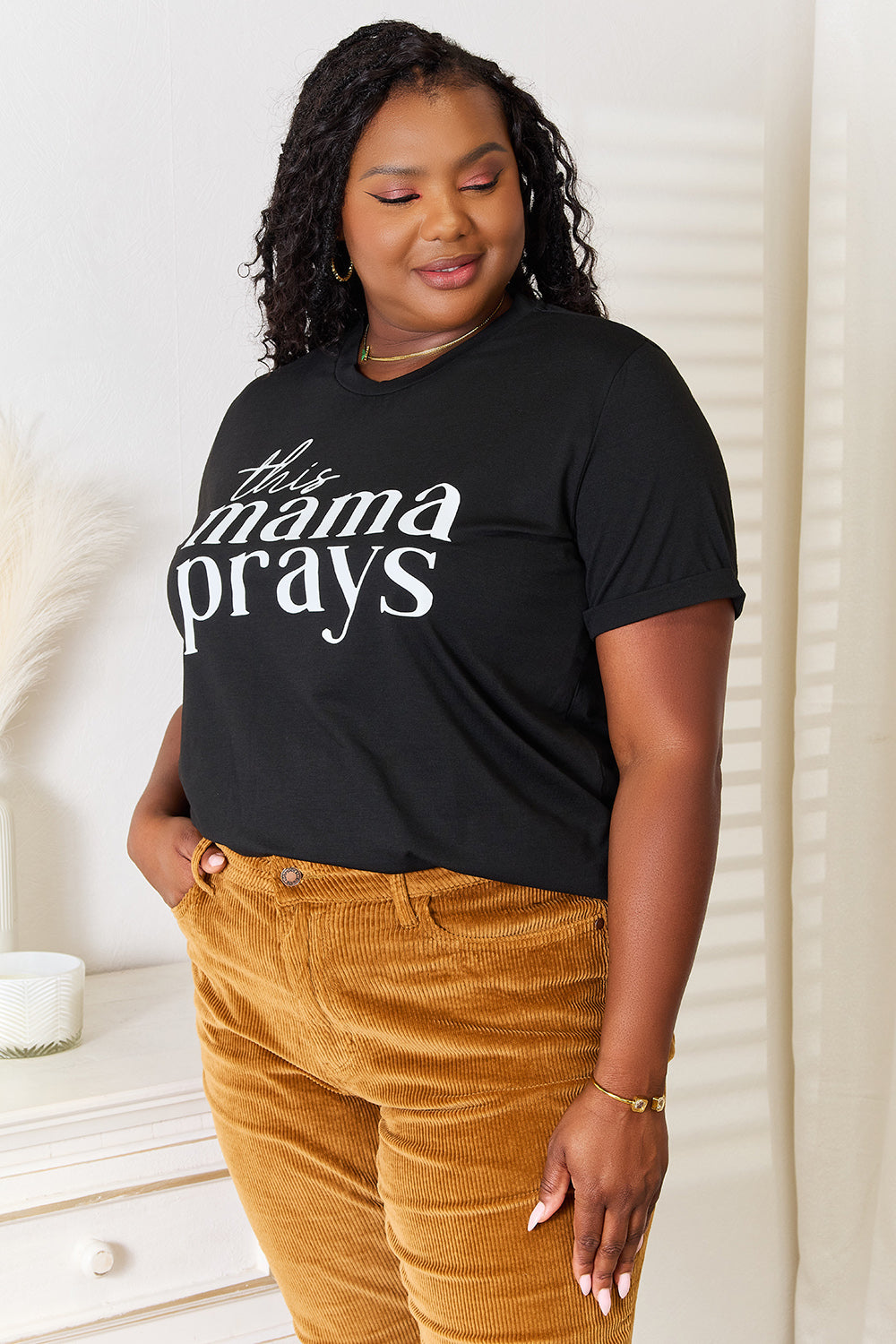 A woman wearing a Simply Love THIS MAMA PRAYS Graphic T-Shirt, paired with brown pants, stands gracefully in a softly lit room, exuding a sense of calm and spiritual gift.