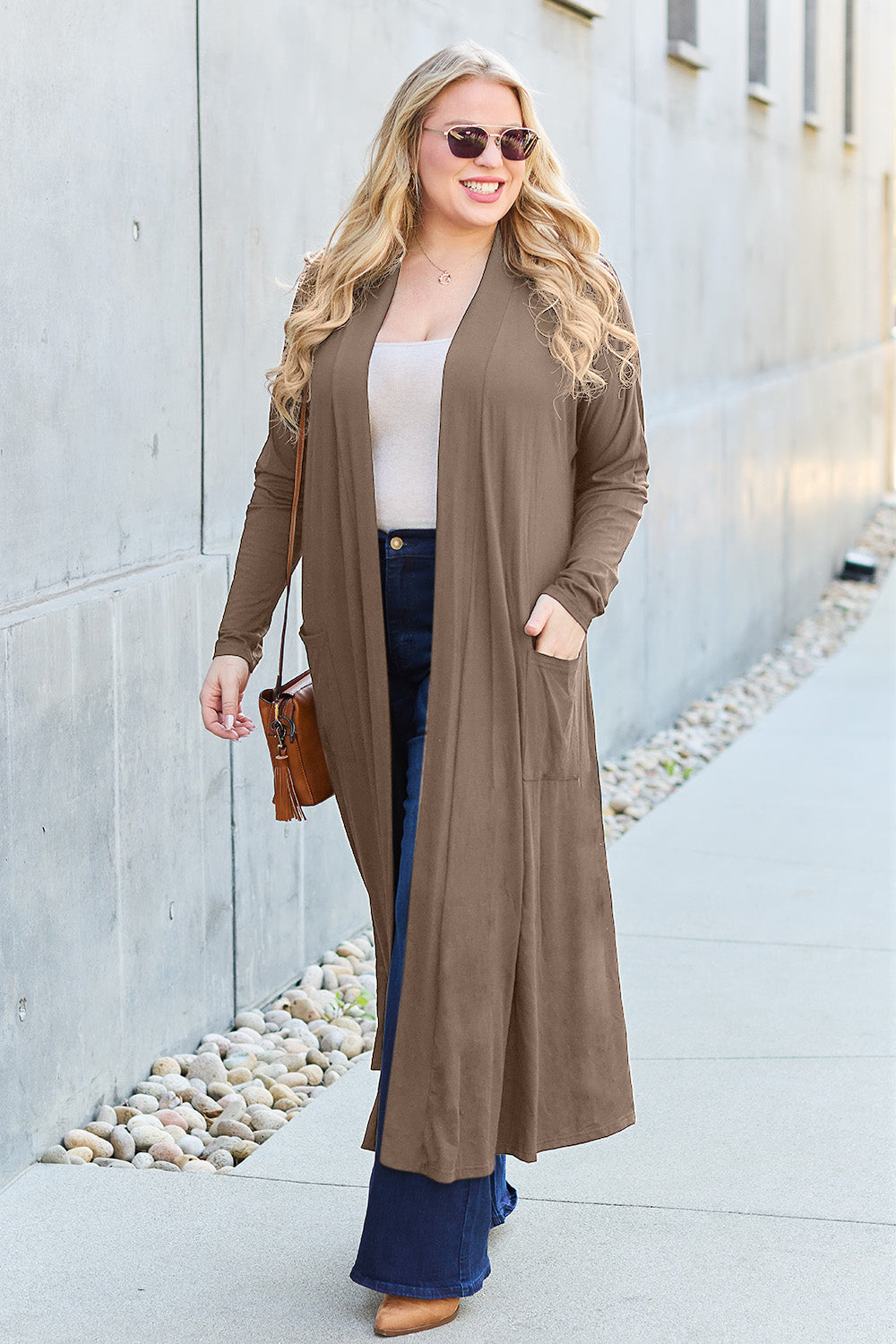 A woman stands against a concrete wall, wearing the Basic Bae Full Size Open Front Long Sleeve Cover Up made from rayon spandex, a white top, blue jeans in her perfect size, and brown shoes with her hands in her pockets. Machine wash cold for best results.