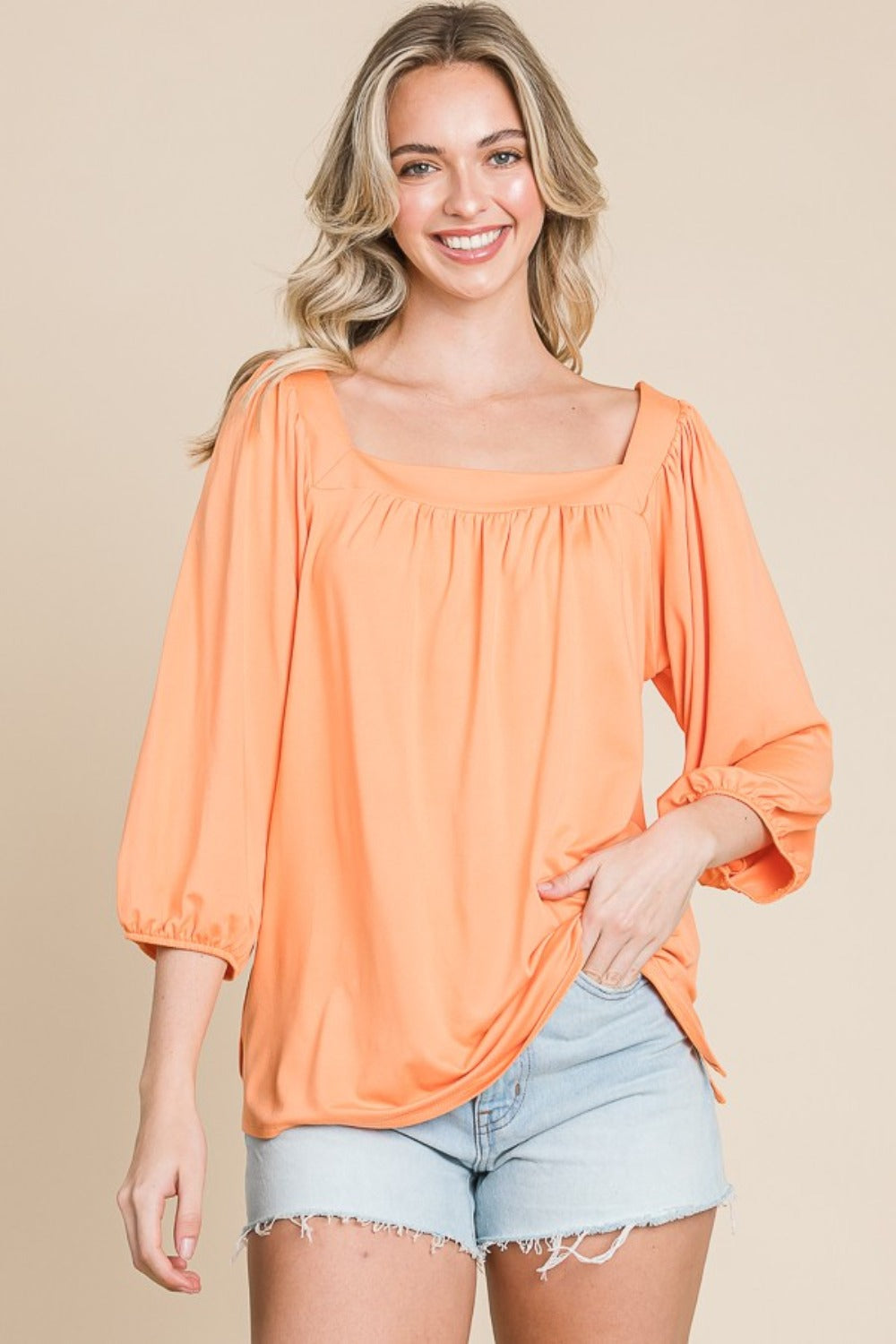 A person with long blonde hair is smiling and wearing the trendy Culture Code Square Neck Puff Sleeve Top in a light orange hue, paired with light denim shorts, showcasing a versatile fashion piece against a beige background.