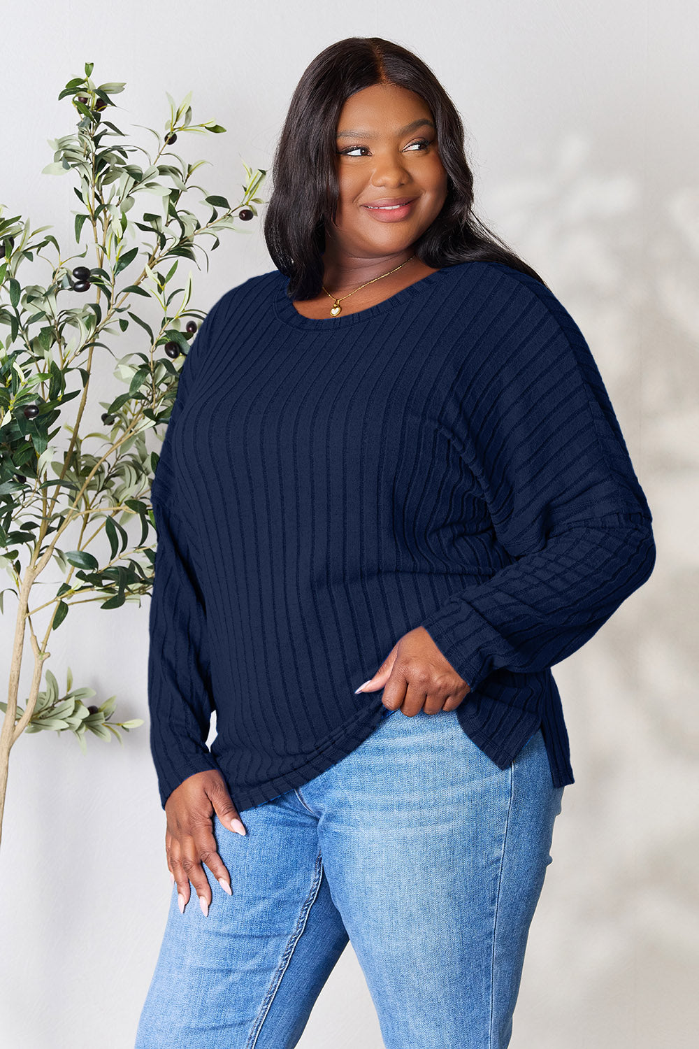 A person wearing the Basic Bae Full Size Ribbed Round Neck Slit Knit Top in blue and beige pants stands and smiles in front of a leafy plant.