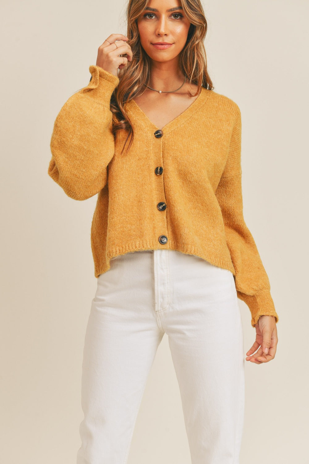 A person wears the MABLE Long Sleeve Button Down Sweater Cardigan in mustard, paired with white high-waisted pants, standing in front of a plain background.