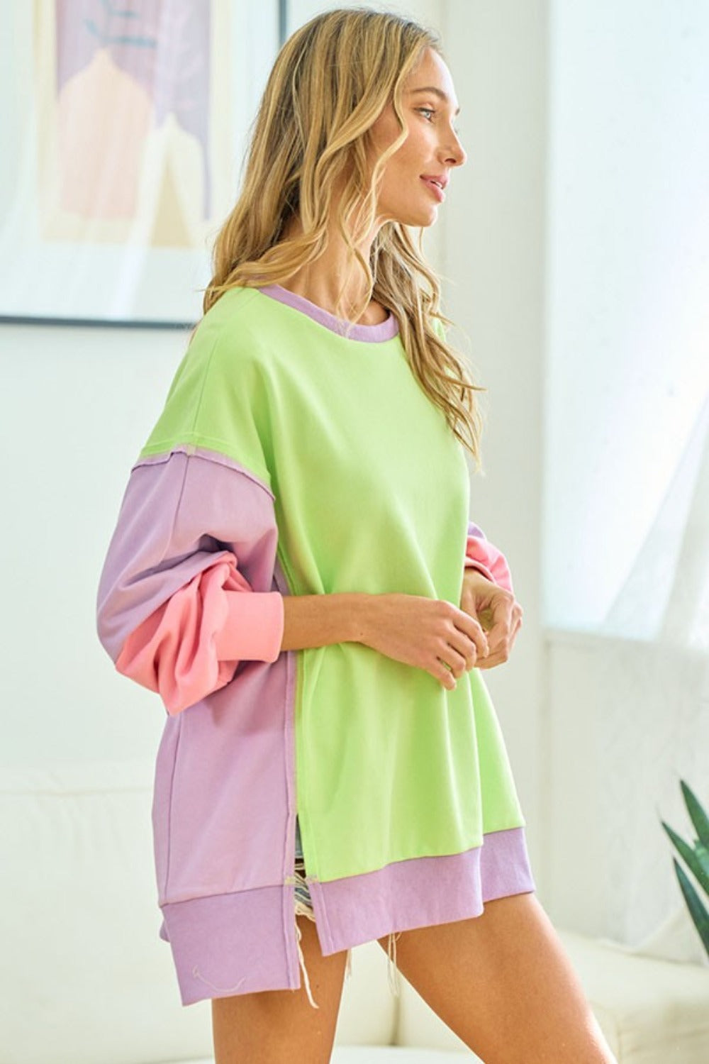 A woman wearing the First Love Color Block Long Sleeve Slit Sweatshirt and matching shorts poses indoors, smiling with one hand touching her hair.