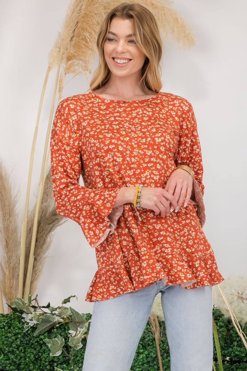 A woman wearing the Celeste Full Size Floral Ruffle Detail Top and jeans stands smiling in front of pampas grass and greenery, exuding feminine flair.