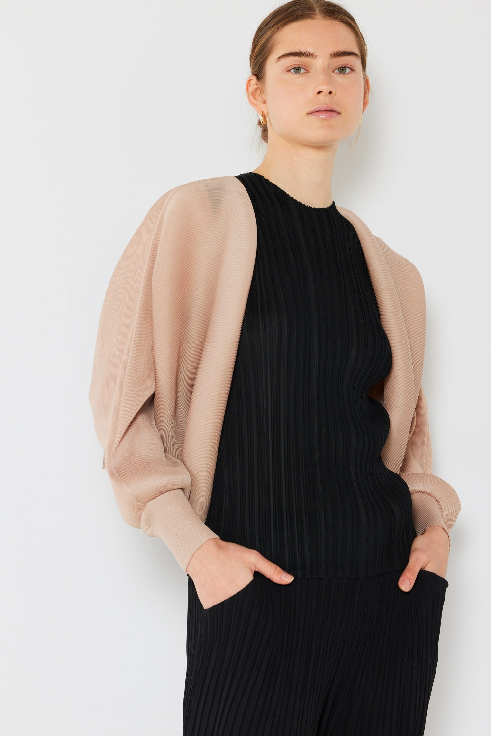 A person stands wearing the stylish Marina West Swim Rib Pleated Puff Sleeve Bolero Cardigan in beige, featuring draped sleeves, paired with a trendy black pleated top.