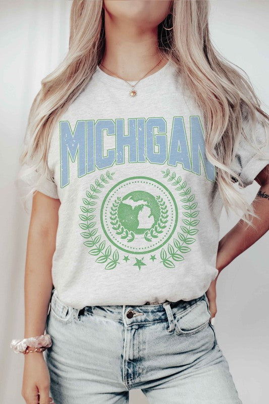 An individual dons a white MICHIGAN Graphic Tee, crafted from 100% cotton. The shirt prominently displays the word "Michigan" in blue, alongside an Earth graphic encircled by laurel leaves and topped with a star. Its unisex sizing guarantees a comfortable fit for all wearers.