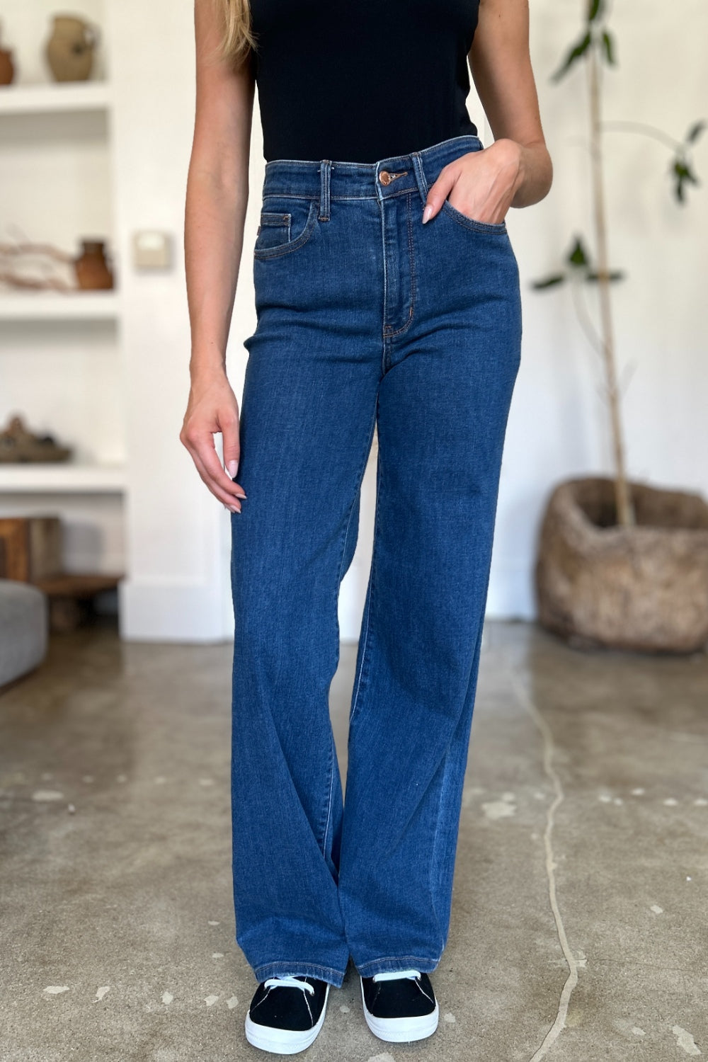 A person wearing Judy Blue Full Size High Rise Straight Jeans, a black top, and white shoes stands gracefully on a polished concrete floor.