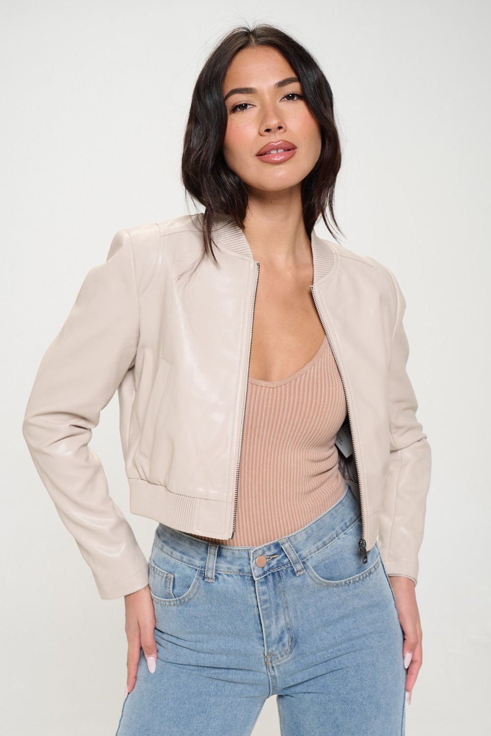 A woman wearing a Coalition LA Zip Up Cropped Bomber Jacket over a ribbed beige top and light blue jeans stands facing the camera, embodying sustainable fashion.