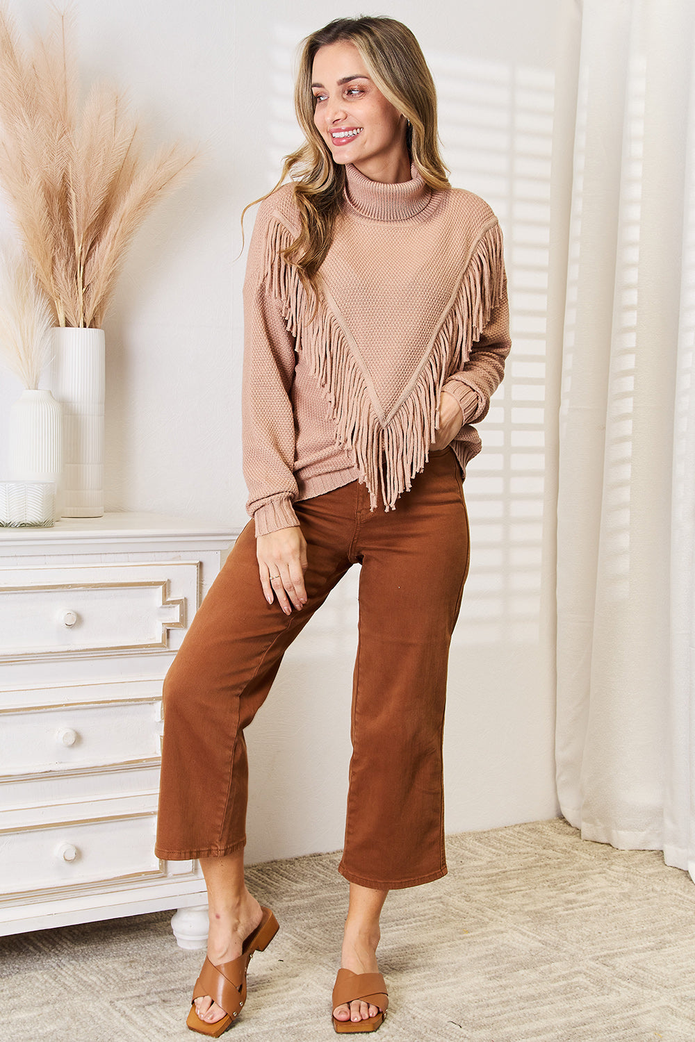 A woman wearing the Woven Right Turtleneck Fringe Front Long Sleeve Sweater, her winter essential, pairs it effortlessly with brown pants. She stands indoors with a neutral expression, embodying cozy sophistication.