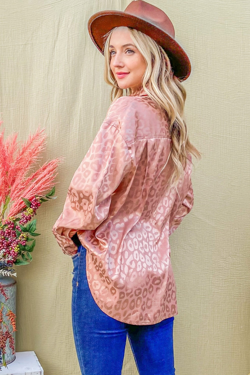 A woman stands in front of a textured background wearing the "And The Why Satin Leopard Button Up Curved Hem Shirt," blue jeans, and a brown hat. She has long hair and is touching the brim of her hat with one hand.