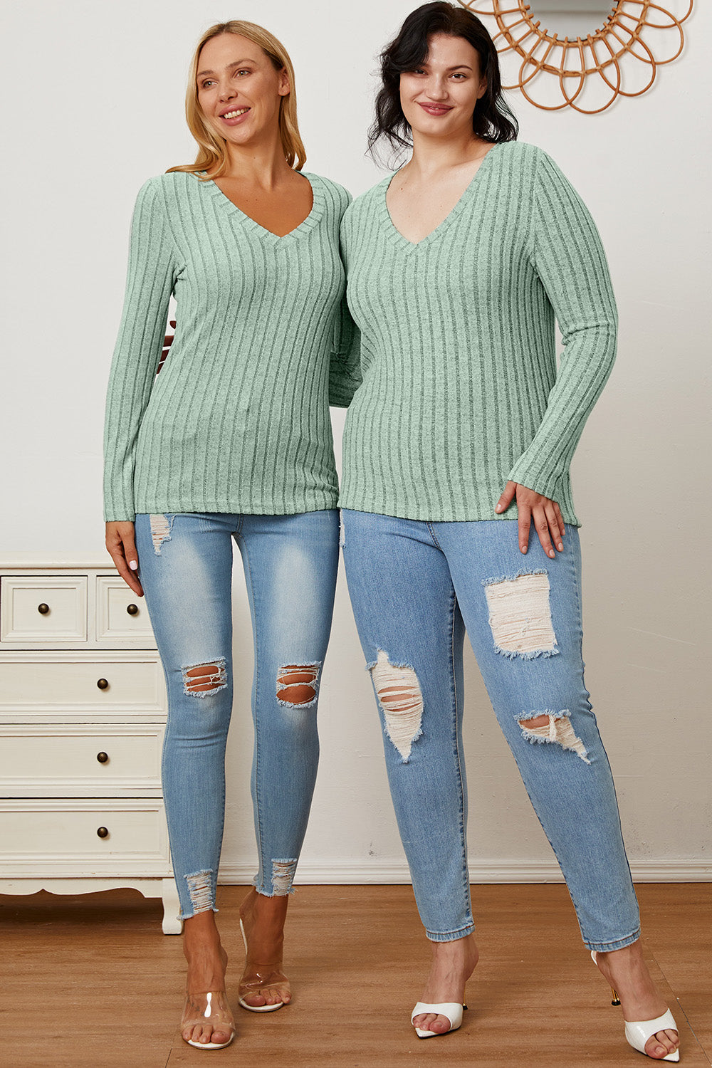 A blonde-haired woman smiles while wearing the Basic Bae Full Size Ribbed V-Neck Long Sleeve T-Shirt, which is light green and slightly stretchy, paired with light blue jeans. She holds one hand near her head and the other near her waist.