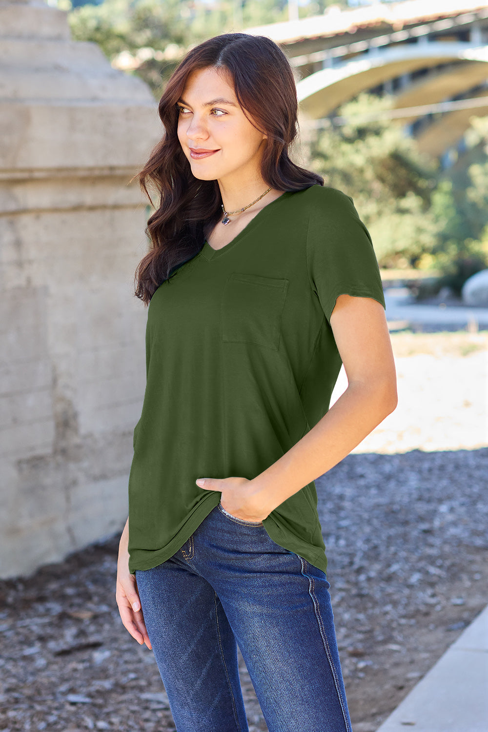 A woman with long brown hair, wearing a Basic Bae Full Size V-Neck Short Sleeve T-Shirt in teal and stretchy blue jeans, stands outdoors near a stone structure, smiling at the camera with one hand in her pocket.