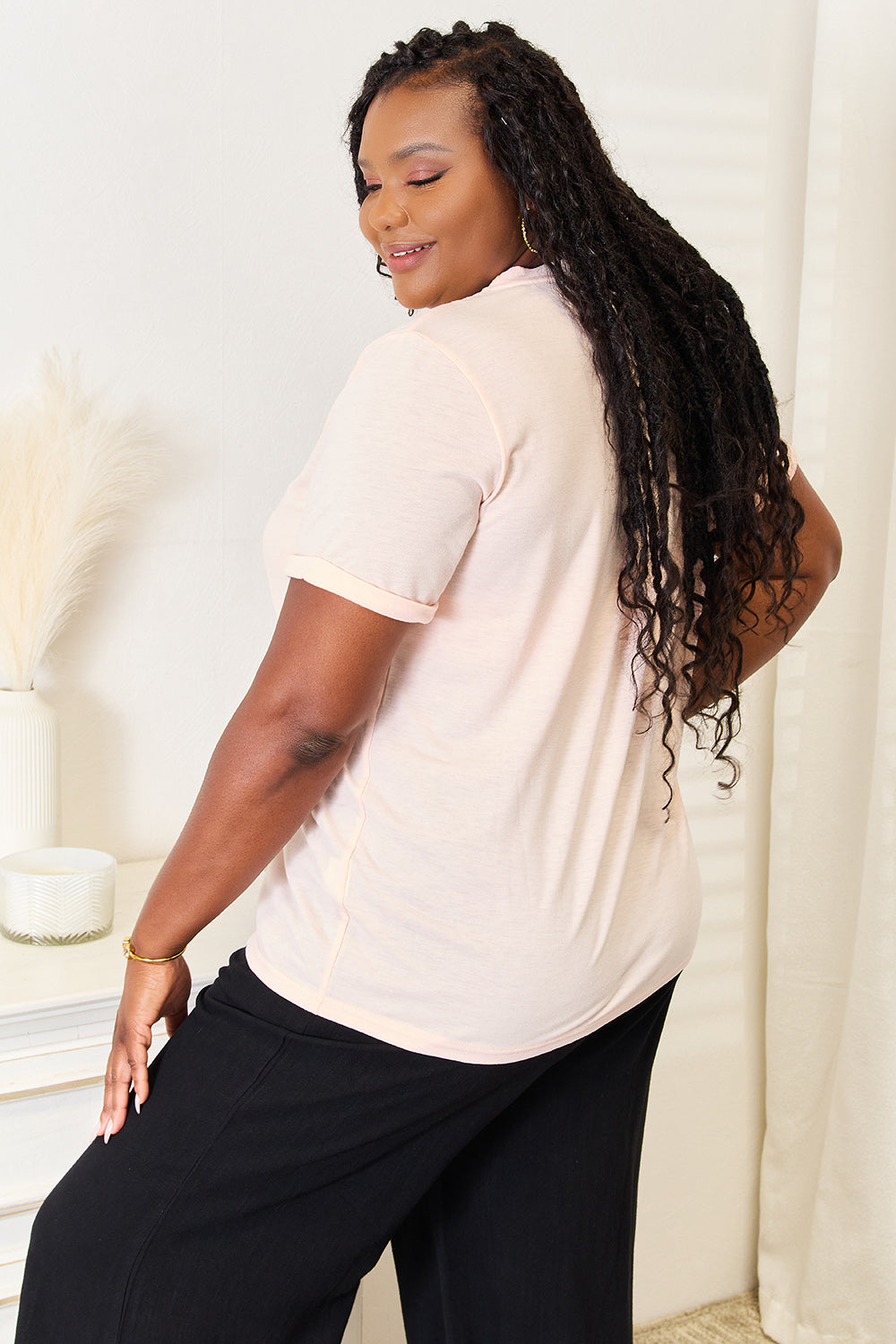 A woman wearing a light pink Simply Love BE KIND Graphic Round Neck T-Shirt with the phrase "be kind" written in cursive. She is standing indoors with a neutral expression, the round neck adding a touch of casual elegance.