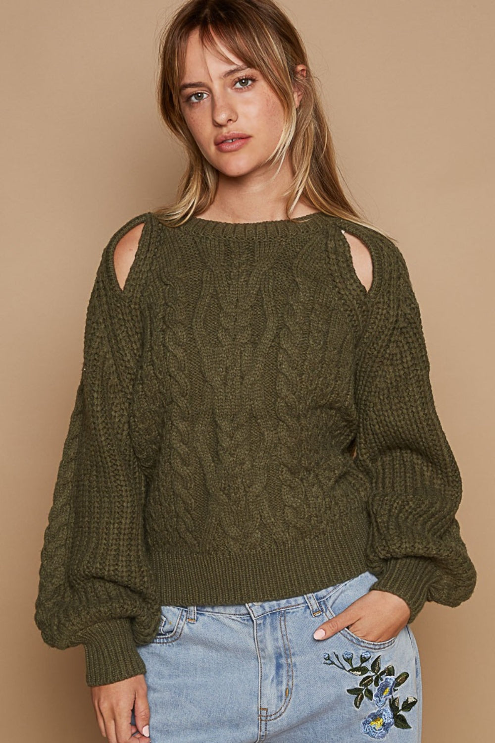 A woman wears the POL Cable Knit Cutout Long Sleeve Sweater in olive-green, showcasing a subtle shoulder keyhole, beautifully matched with light blue jeans adorned with floral embroidery, all set against a beige background.