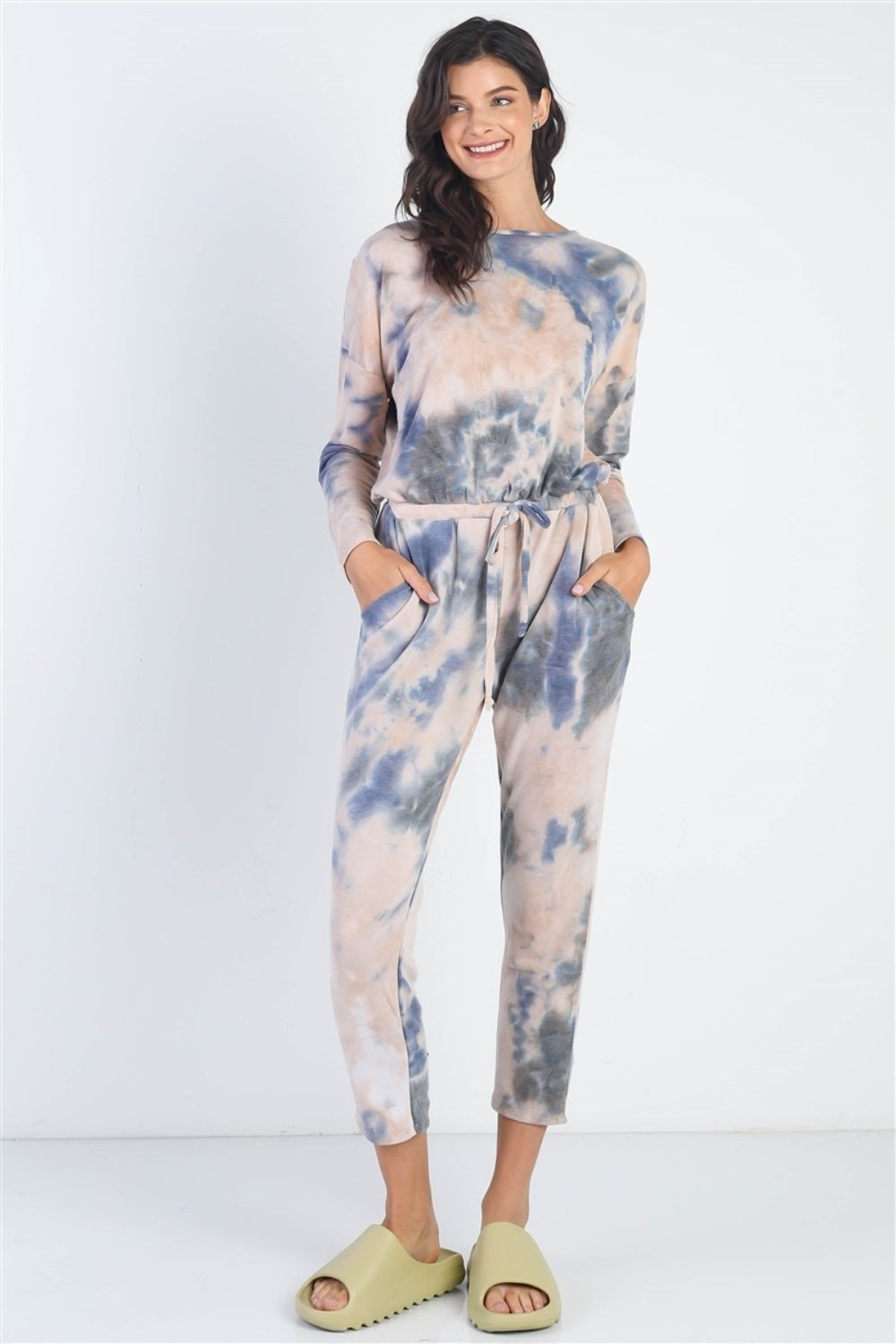 Dressed in a Cherish Apparel Tie-Dye Round Neck Long Sleeve Jumpsuit and green sandals, a person strikes a street-style pose against a plain white background.