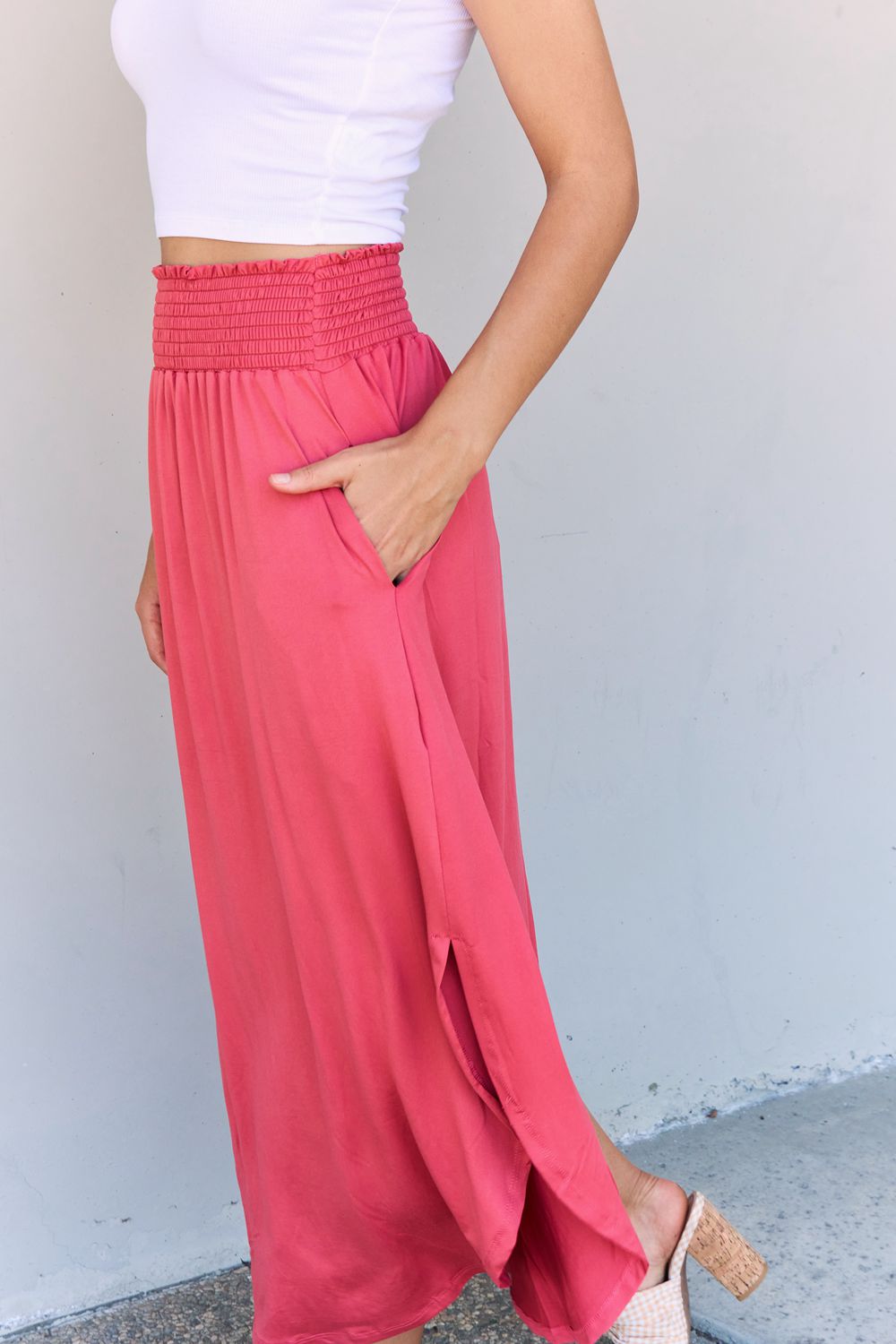 A person wearing a white top and the Doublju Comfort Princess Full Size High Waist Scoop Hem Maxi Skirt in an elegant coral color, paired with tan sandals, stands in front of a plain wall.