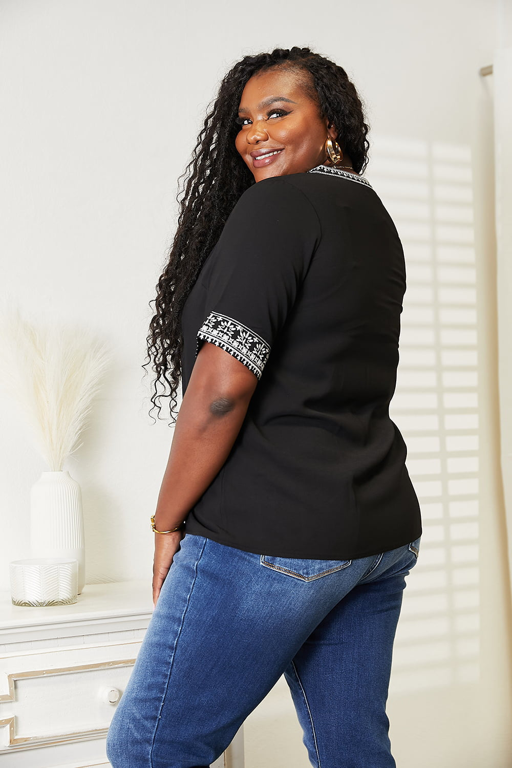 Wearing the elegant Double Take Embroidered Notched Neck Top and blue jeans, a woman with long, wavy hair stands smiling against a light background.