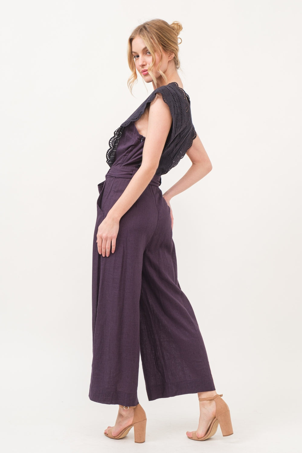 A person in an elegant and fashionable And The Why Laced Surplice Tie Waist Jumpsuit with wide-leg pants stands against a plain background, smiling with hands on hips.