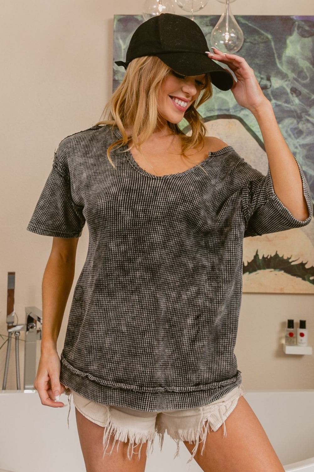 Woman wearing a black cap, the BiBi Washed Notched Short Sleeve Top in a loose-fitting gray wash, and distressed white shorts, smiling and holding the brim of her hat while standing indoors. This versatile casual piece can be effortlessly layered under a jacket for a chic look.