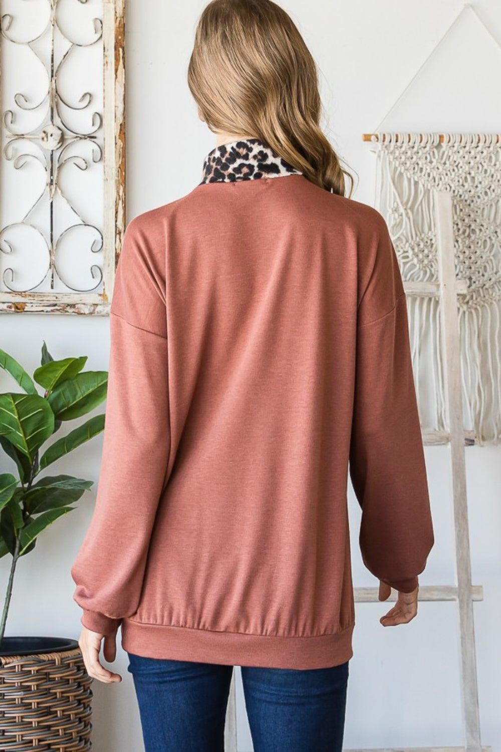 A woman wearing the stylish Heimish Full Size Leopard Quarter Zip Mock Neck Sweatshirt stands indoors near a lush plant and an intricate macrame wall hanging.