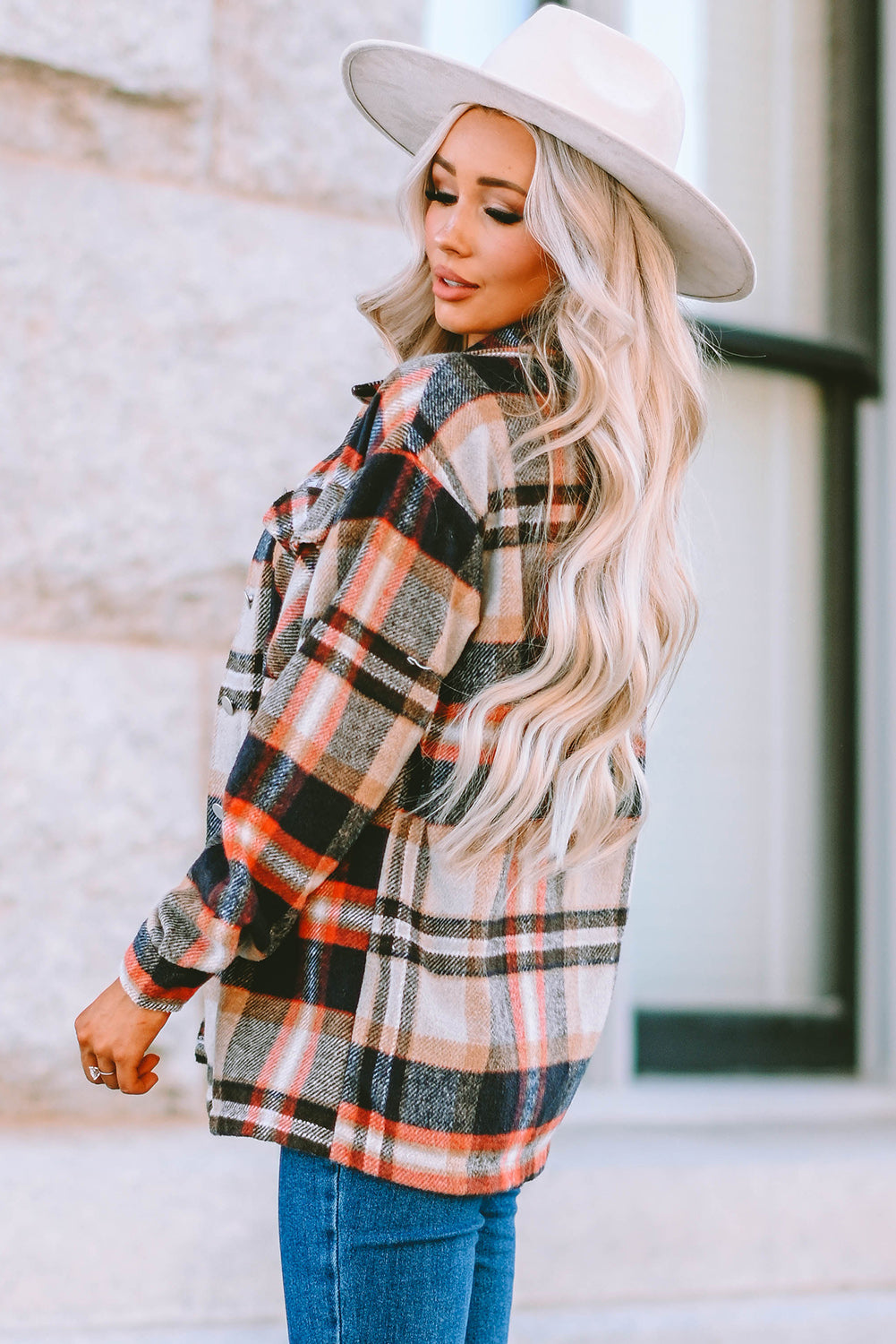 A person wearing a Brown Plaid Button Up Long Sleeve Flannel Shacket layered over a white top with ripped jeans and holding a brown handbag stands against a textured gray wall.