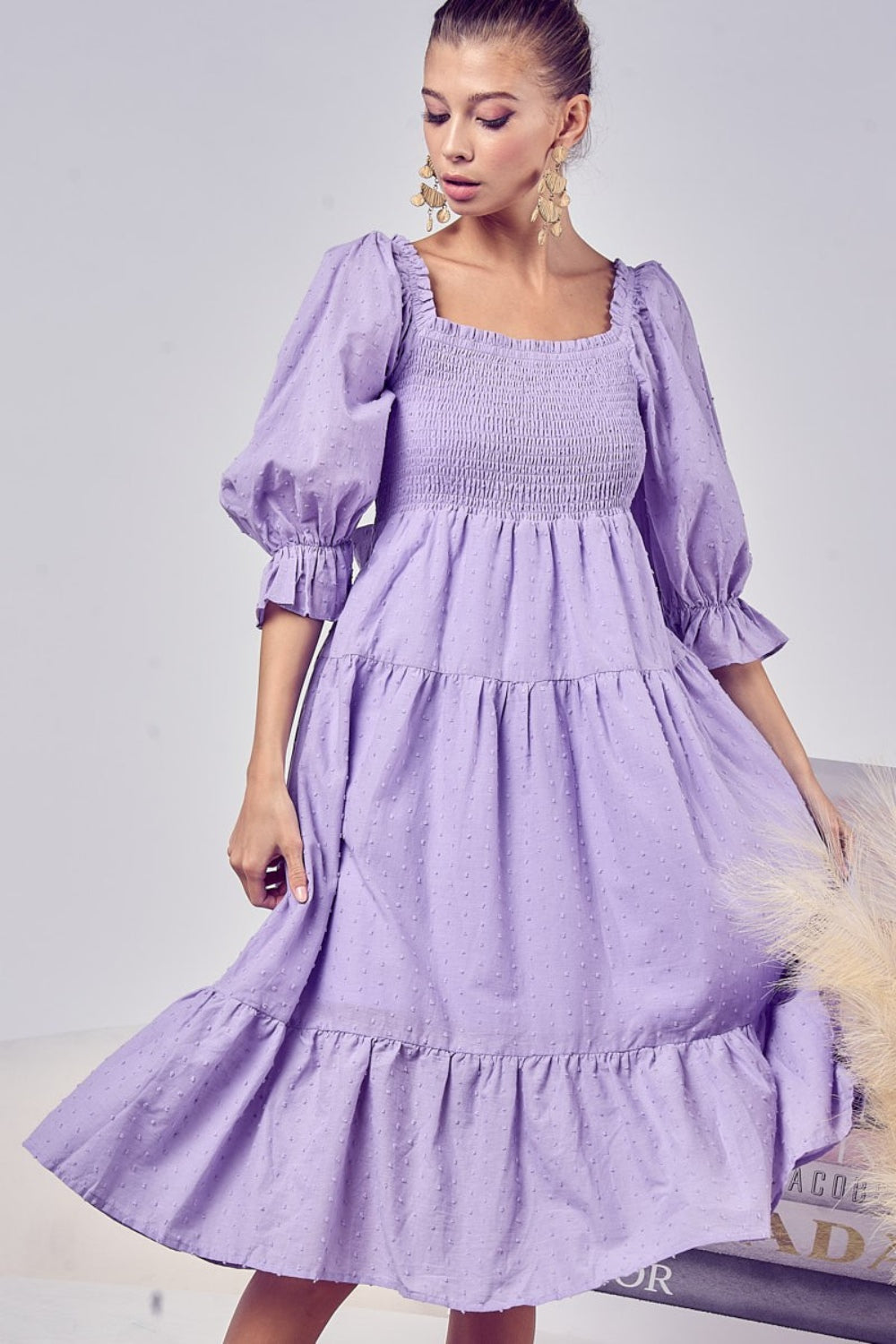 A woman stands smiling in the BiBi Swiss Dot Flounce Sleeve Smocked Tiered Midi Dress, showcasing its lavender hue, long flounce sleeves, tiered skirt, and delicate Swiss dots.