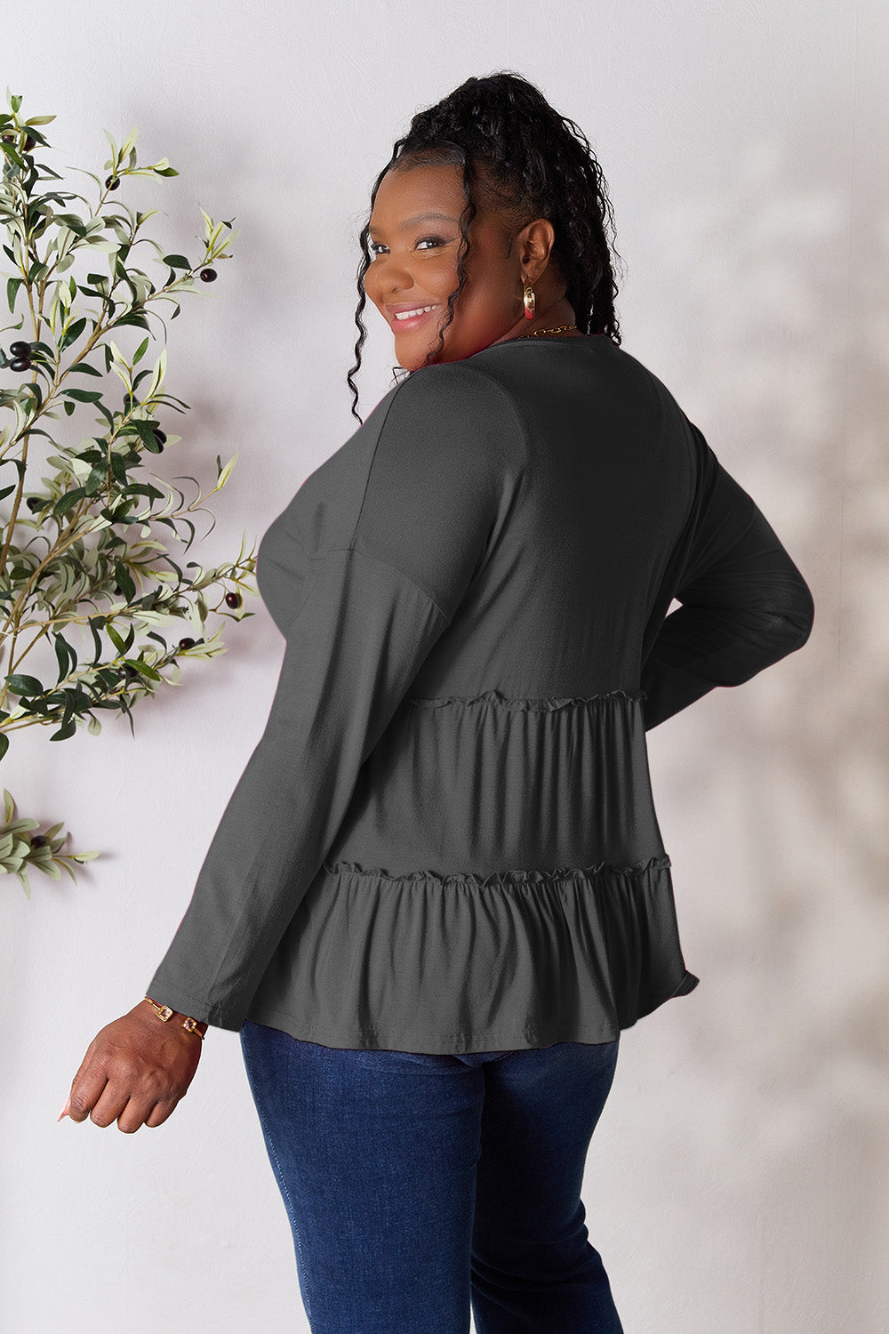 Person stands indoors near a plant, wearing the Double Take Half Button Long Sleeve Ruffle Hem Blouse in gray and blue jeans, smiling while looking to the side. The slightly stretchy fabric of the blouse adds comfort while its buttoned design offers a touch of elegance.