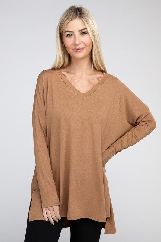 A woman with long, blonde hair is wearing a loose-fitting, brown Dolman Long Sleeve V-Neck Side Slit Hi-Low Hem Top and black pants. She poses with one hand on her hip and a neutral facial expression against a light grey background.