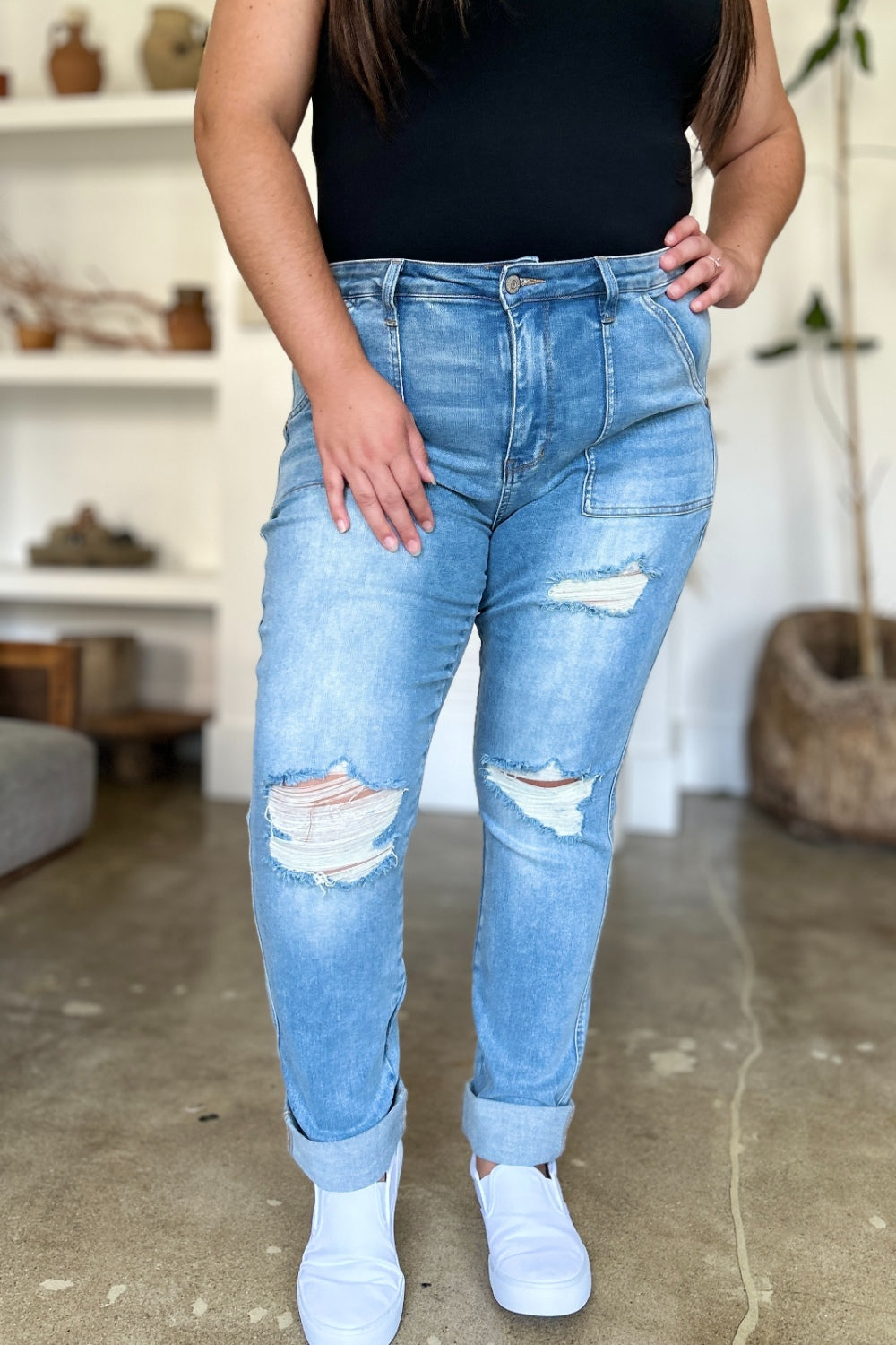 Two women are standing side by side, both clad in black and white tank tops and Judy Blue Full Size Distressed Straight Jeans with Patch Pockets, smiling at the camera in a modern living room.