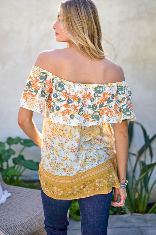 A woman with blonde hair is smiling while standing outdoors, wearing the Printed Off Shoulder Smocked Top paired with jeans.