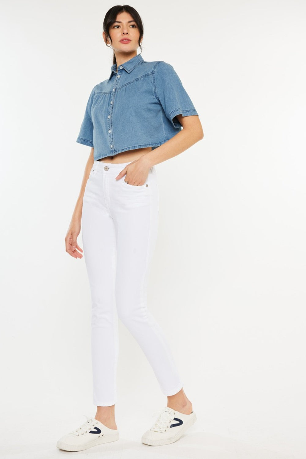 A person wearing a short-sleeve denim button-up shirt, Kancan High Rise Ankle Skinny Jeans in classic white, and white sneakers stands effortlessly against a plain background.