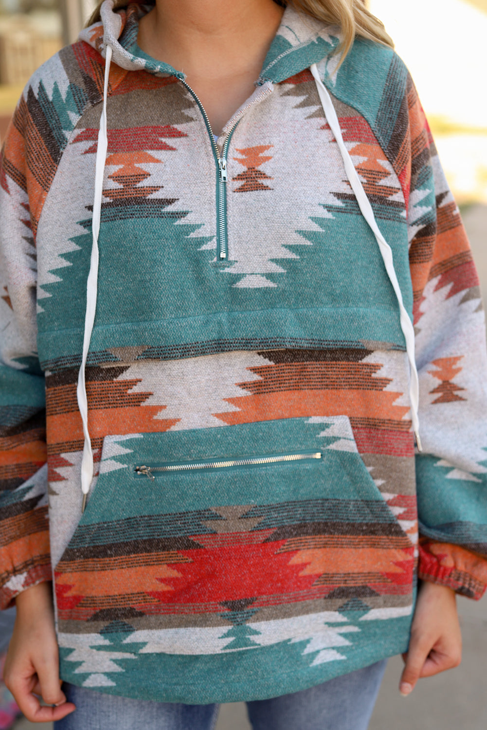 A woman with long blonde hair is standing outside, facing away from the camera. She wears a Multicolor Aztec Print Zipped Split Neck Hoodie and jeans. A rug and potted plant are visible in the background, adding to the cozy vibe of her super comfortable outfit.