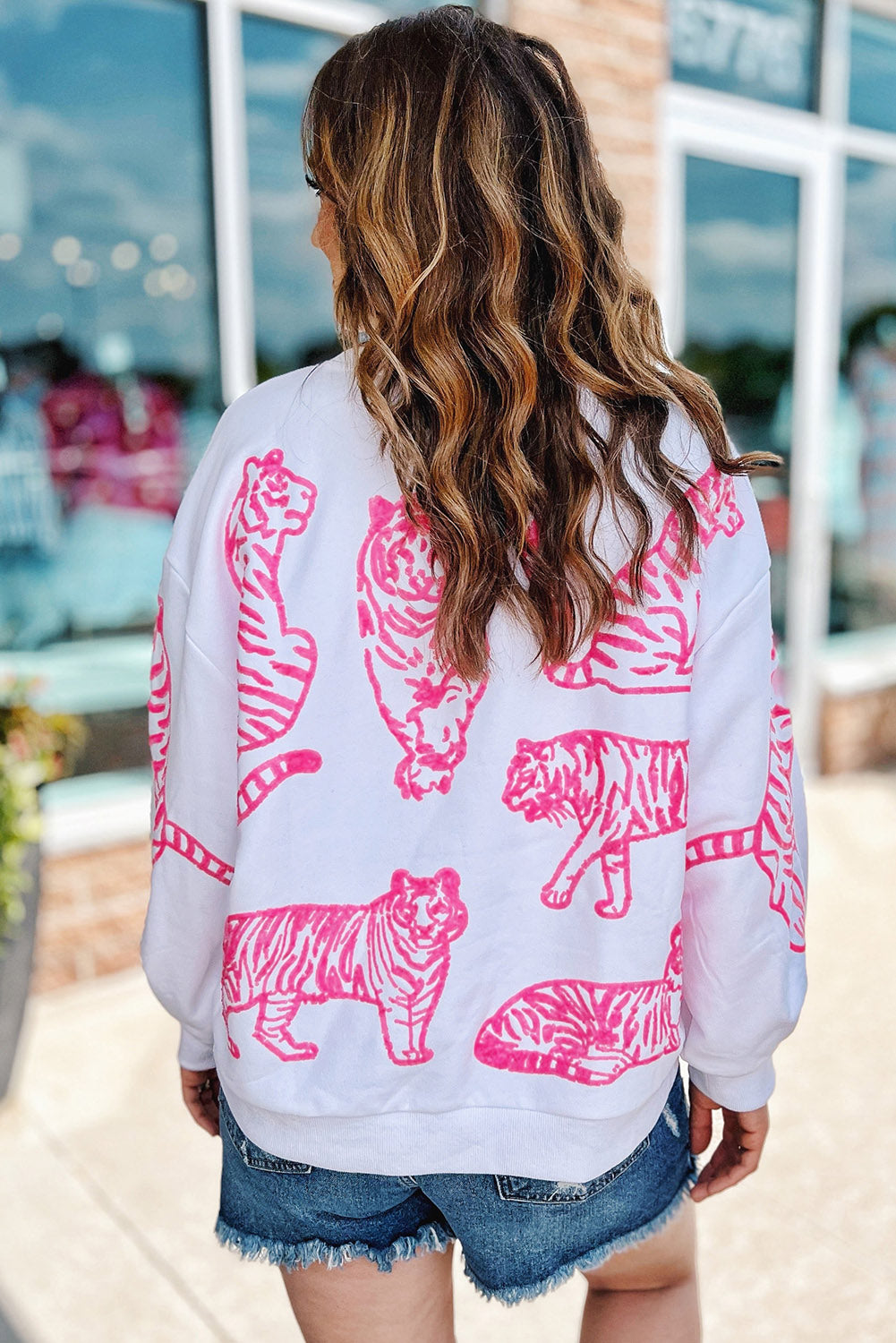 A person with long blonde hair wears a trendy Bright White Pink Animal Print Pullover Sweatshirt, paired with blue jeans, standing outdoors with their back facing the camera.