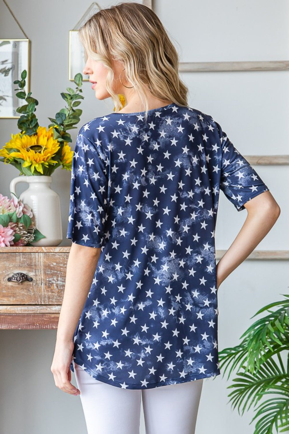 A person with long blonde hair is wearing the Heimish Full Size Short Sleeve Crisscross Star Babydoll Top in blue and white pants, standing indoors near a table adorned with flowers and framed pictures on the wall.