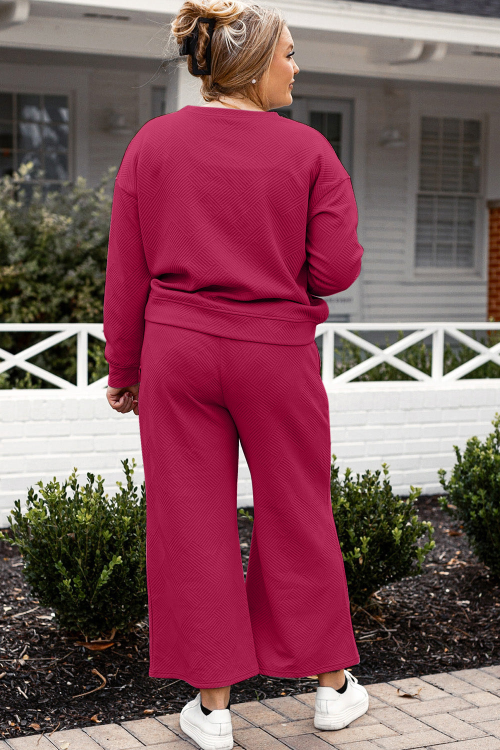 A woman with blonde hair, dressed in the Double Take Full Size Textured Long Sleeve Top and Drawstring Pants Set featuring a stretchy black long-sleeve top and matching wide-leg trousers made of rayon spandex, stands barefoot in a room with white walls and a wooden floor.