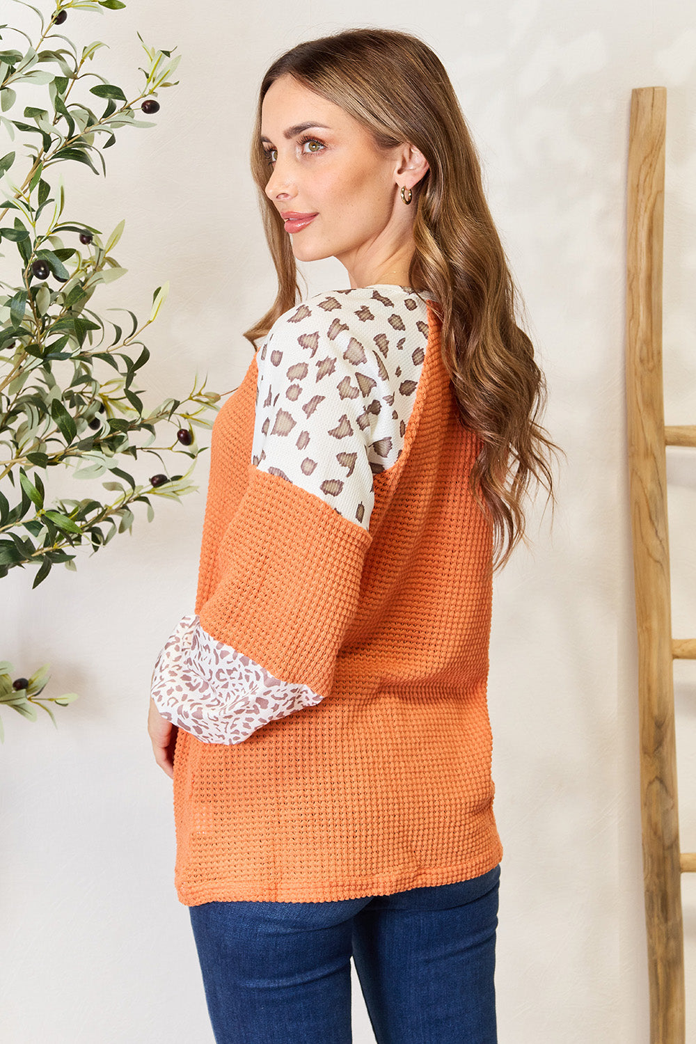 A woman poses, smiling, wearing the trendy Leopard Waffle-Knit Long Sleeve Blouse paired with blue jeans, standing near a wooden ladder and a plant.