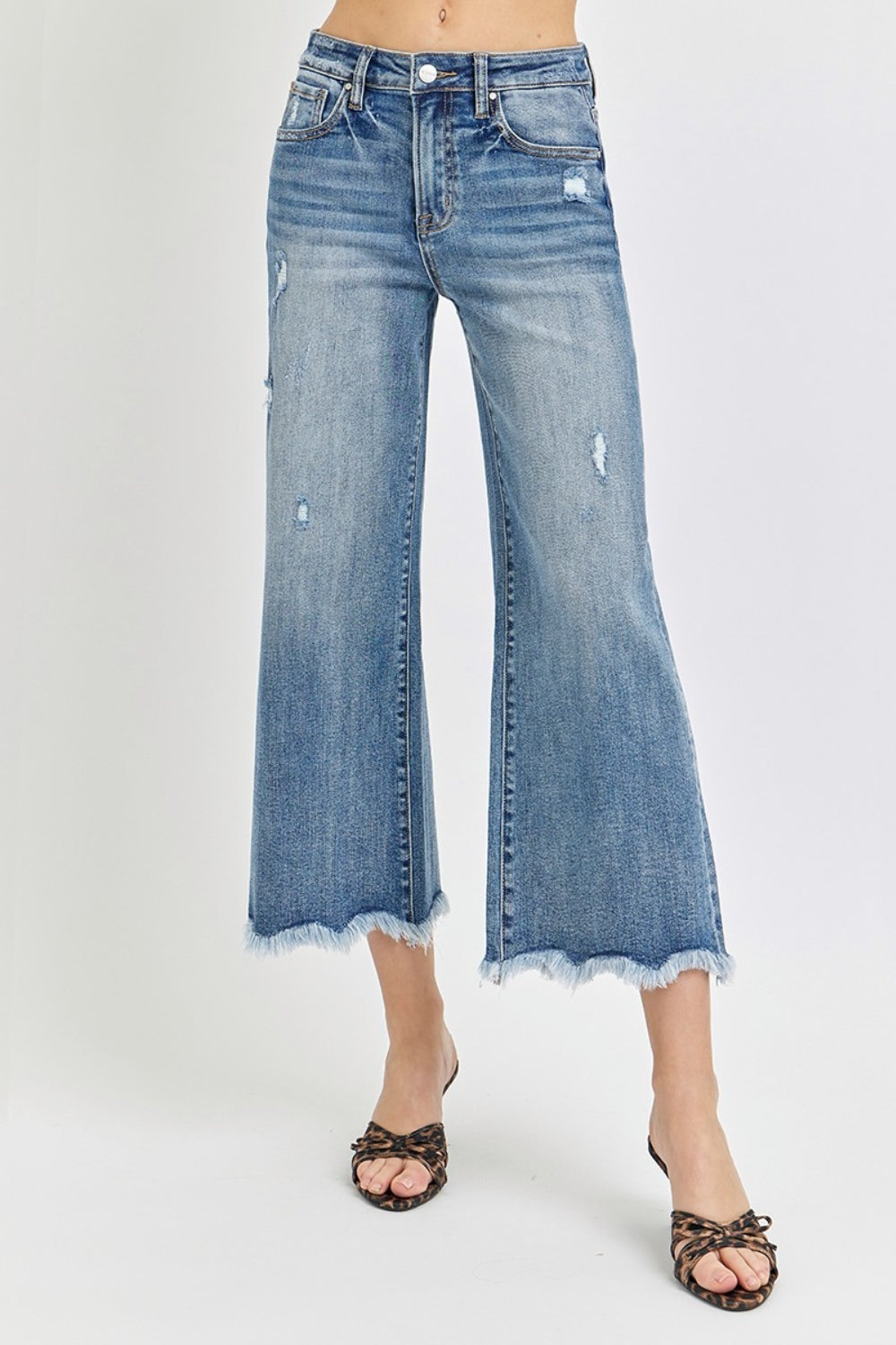 Person wearing Risen High Rise Cropped Flare Jeans with frayed details and a few rips, paired with strappy high-heeled sandals against a plain background.