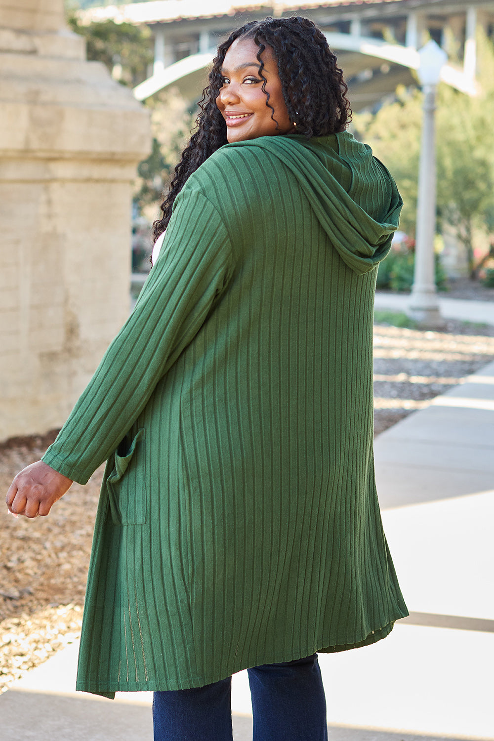 A woman wearing the Basic Bae Full Size Hooded Sweater Cardigan in black, paired with a white top and khaki pants, stands in front of a light background accented by a leafy plant on her left. With one hand tucked into her pocket, she gazes to her right.