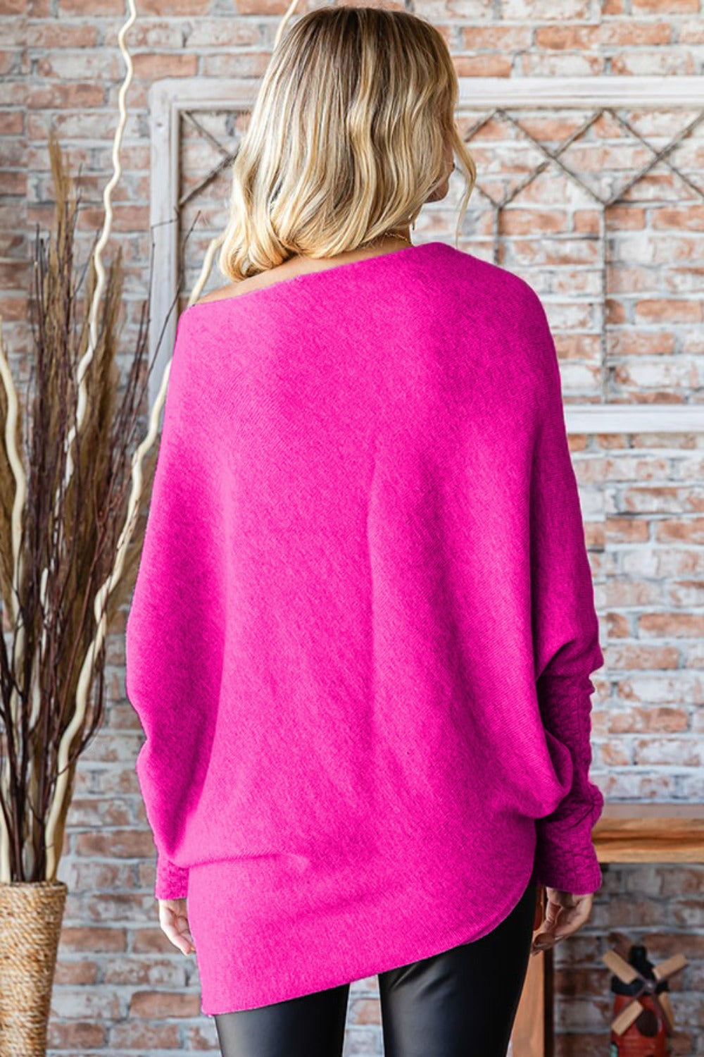 A person with long, blonde hair is standing indoors, smiling, and wearing a bright pink First Love Full Size Asymmetrical Hem Dolman Sleeve Sweater paired with black pants.