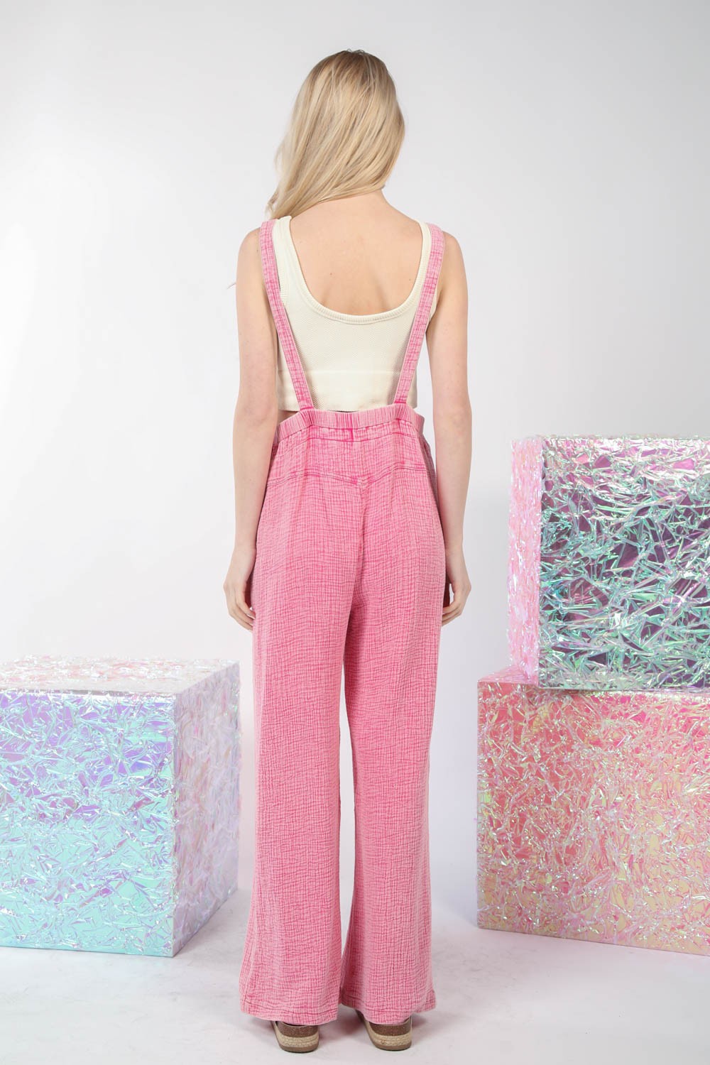 The person is wearing VERY J Texture Washed Wide Leg Overalls in pink with a vintage charm and sandals, standing next to pastel-colored reflective cubes against a white background.