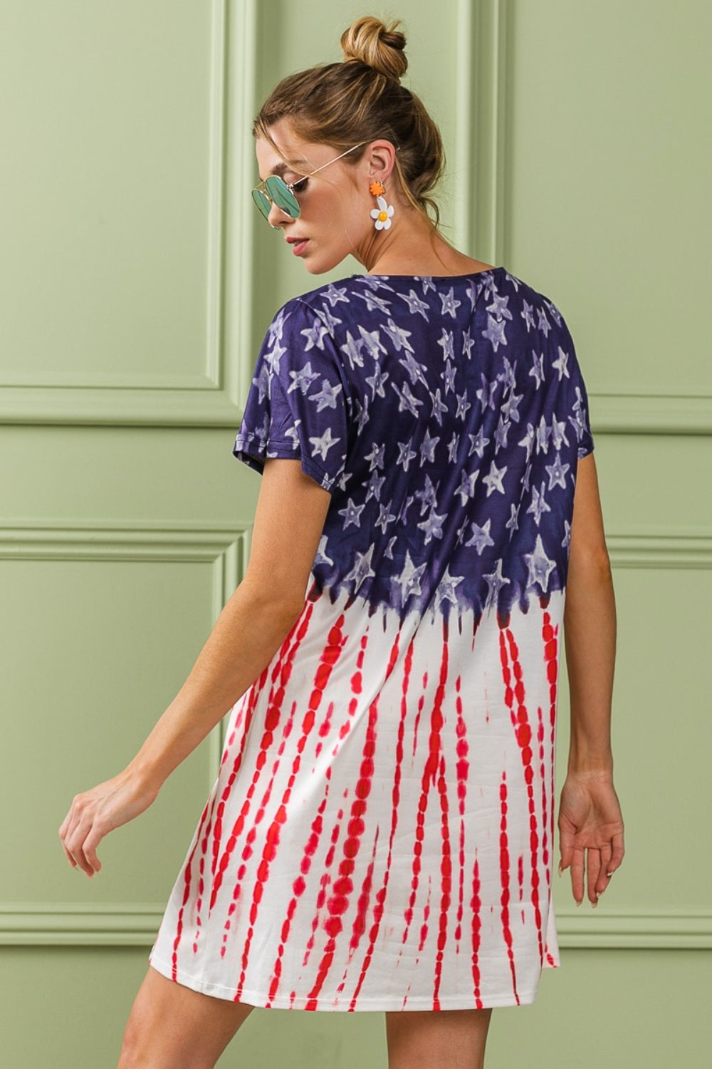 A person wearing the BiBi American Flag Theme Tee Dress, accessorized with sunglasses and earrings, stands against a light green wall with decorative molding.