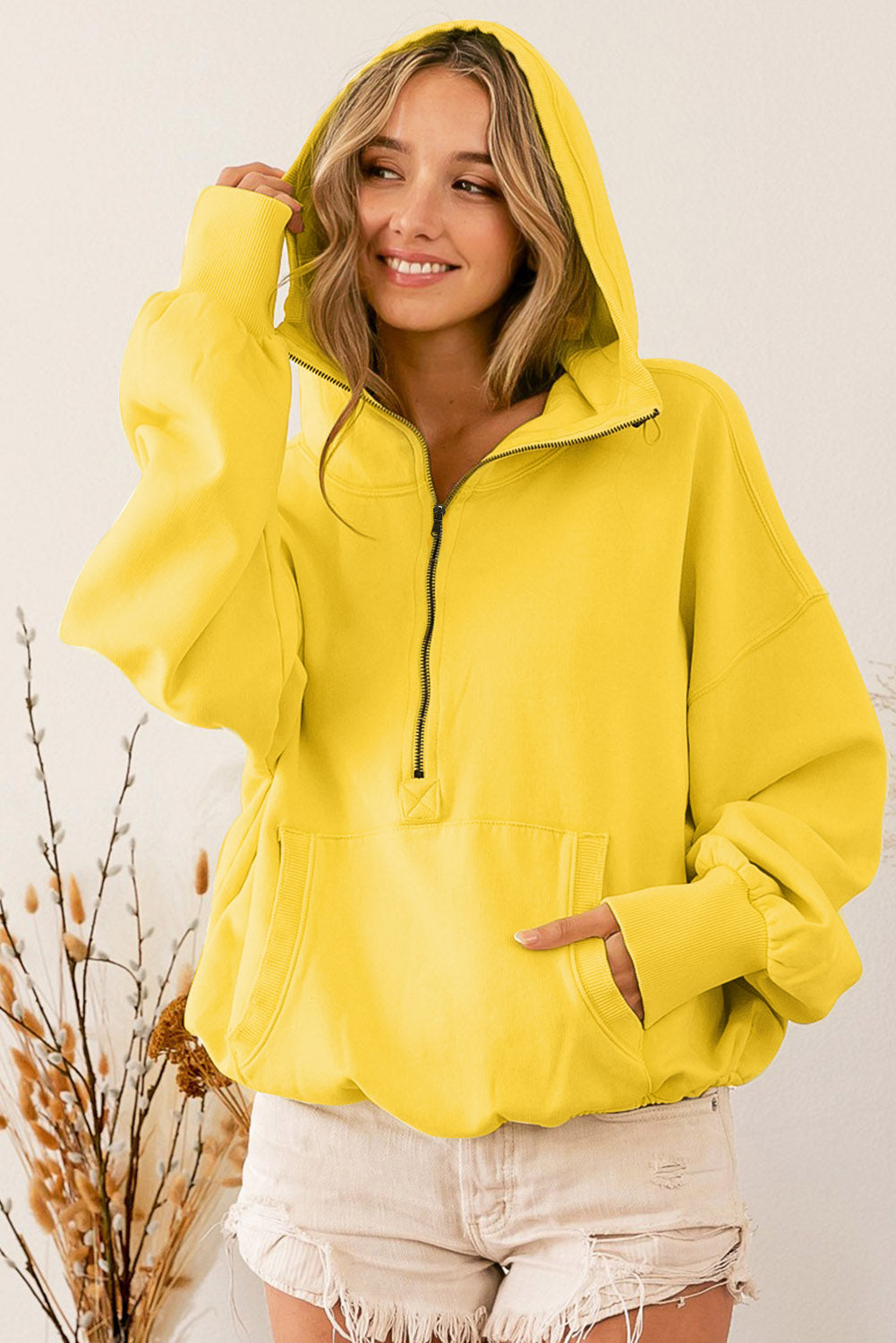 A person with long hair is wearing a Yellow Ribbed Trim Kangaroo Pocket Zipped Hoodie and frayed beige shorts, standing sideways in a room decorated with dried plants.