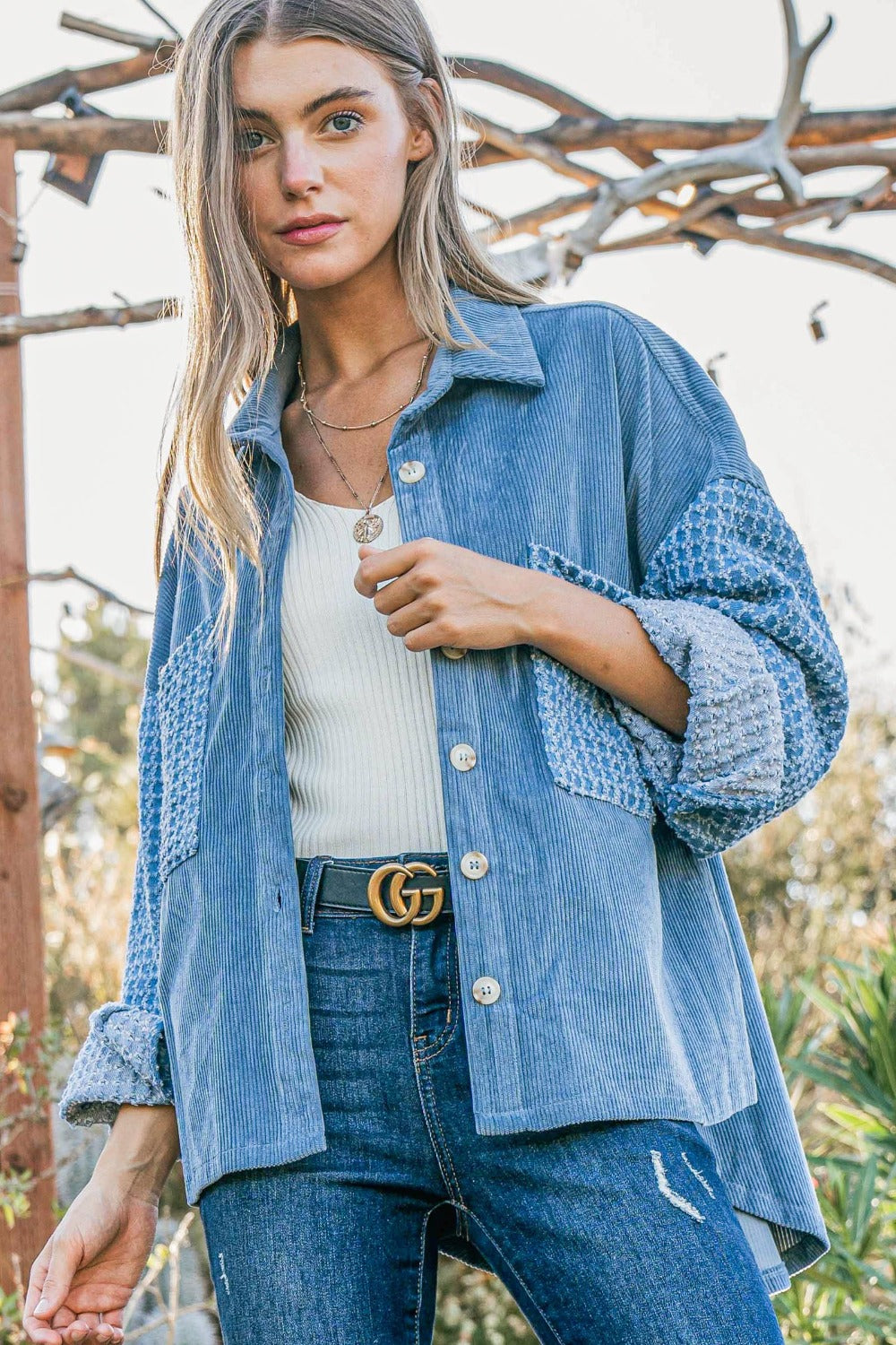 A woman stands outdoors wearing the And The Why Button Down Corduroy Contrast Texture Long Sleeve Shacket over a white top and blue jeans. She has long hair and wears a belt with a prominent buckle.