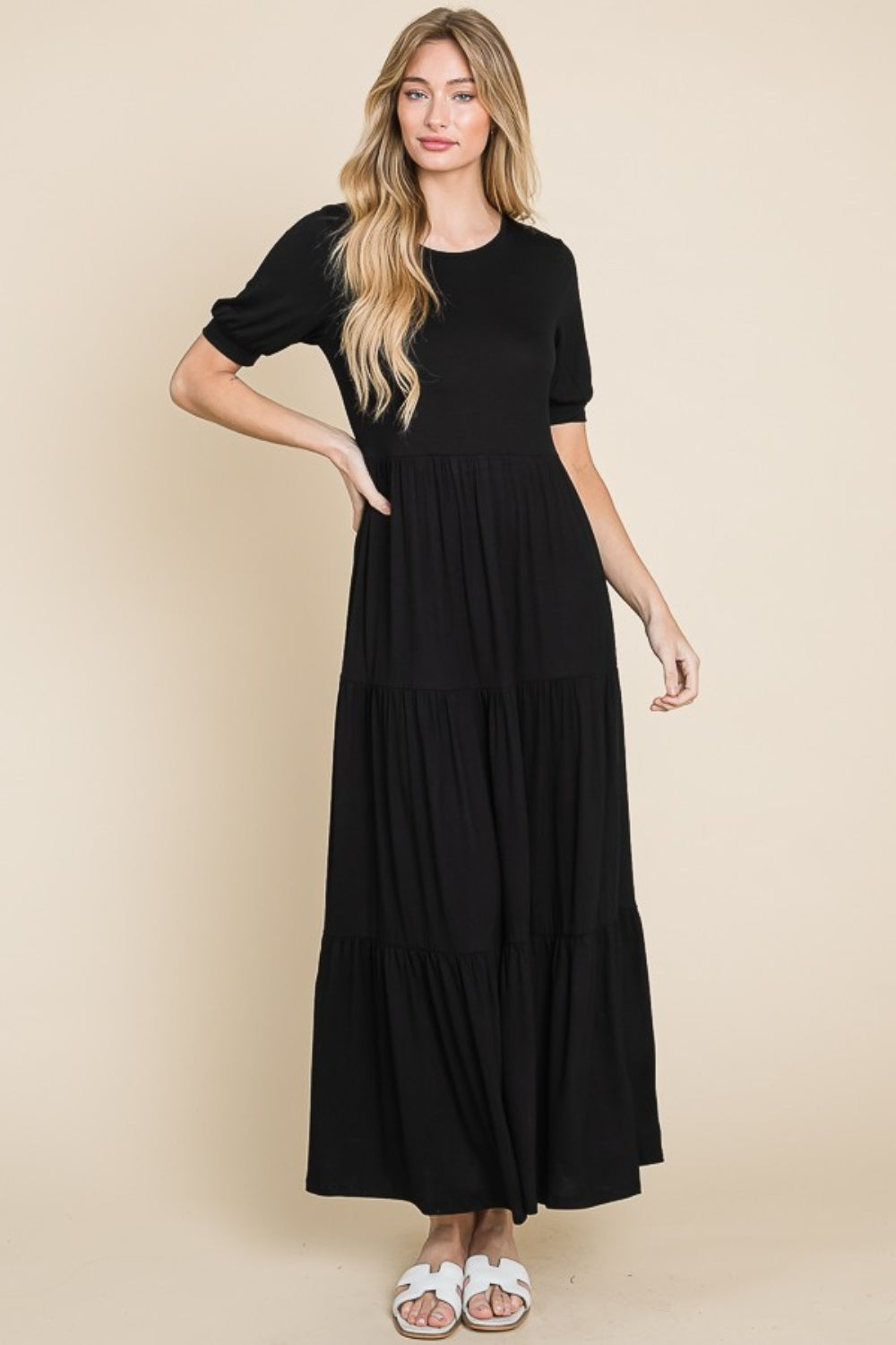 A person wearing the BOMBOM Short Sleeve Tiered Maxi Dress in sleek black stands against a plain beige background, exuding comfort with style. With one hand on their hip and donning white sandals, they effortlessly showcase this versatile piece.