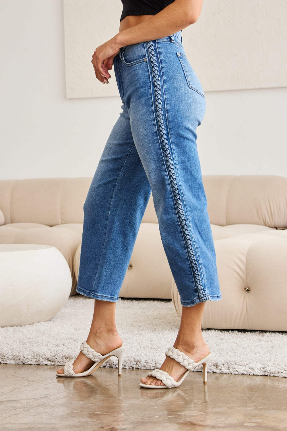 A person wearing Judy Blue Full Size Braid Side Detail Wide Leg Jeans and black heels is standing on a polished floor. There is a white couch and a large potted plant in the background.