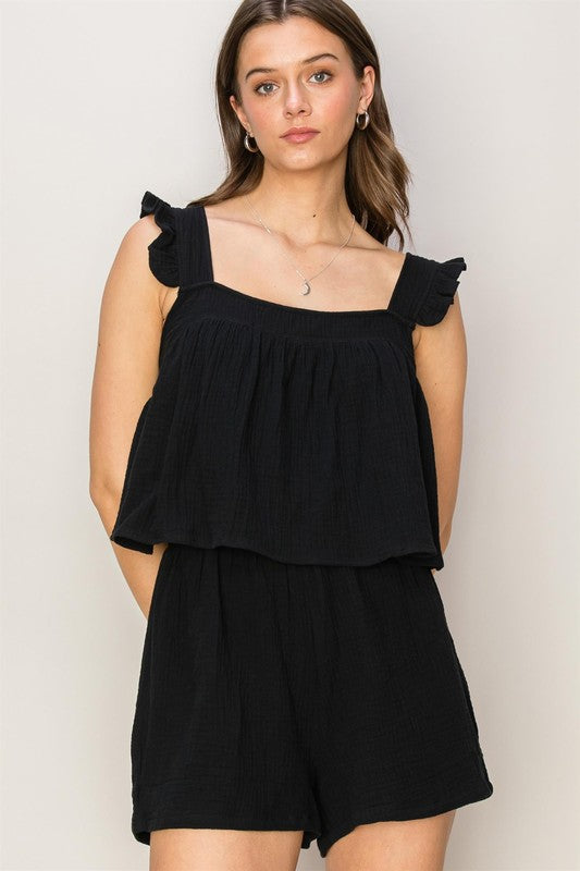 A person stands against a plain background, wearing the Double Gauze Top and Shorts Set, featuring a sleeveless black romper with ruffled straps and a stylish square neck.