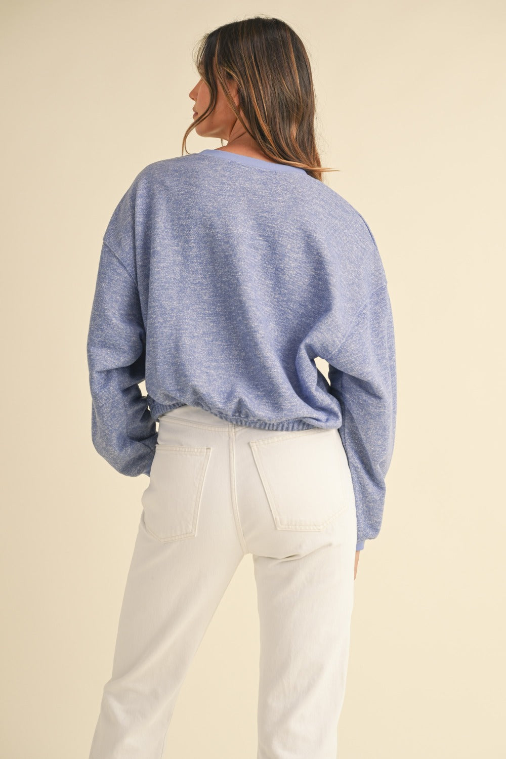 A person wearing a Mable Round Neck Letter Embroidery Crop Sweatshirt in blue, paired with white pants, stands against a plain background, touching the collar of the sweatshirt with one hand.