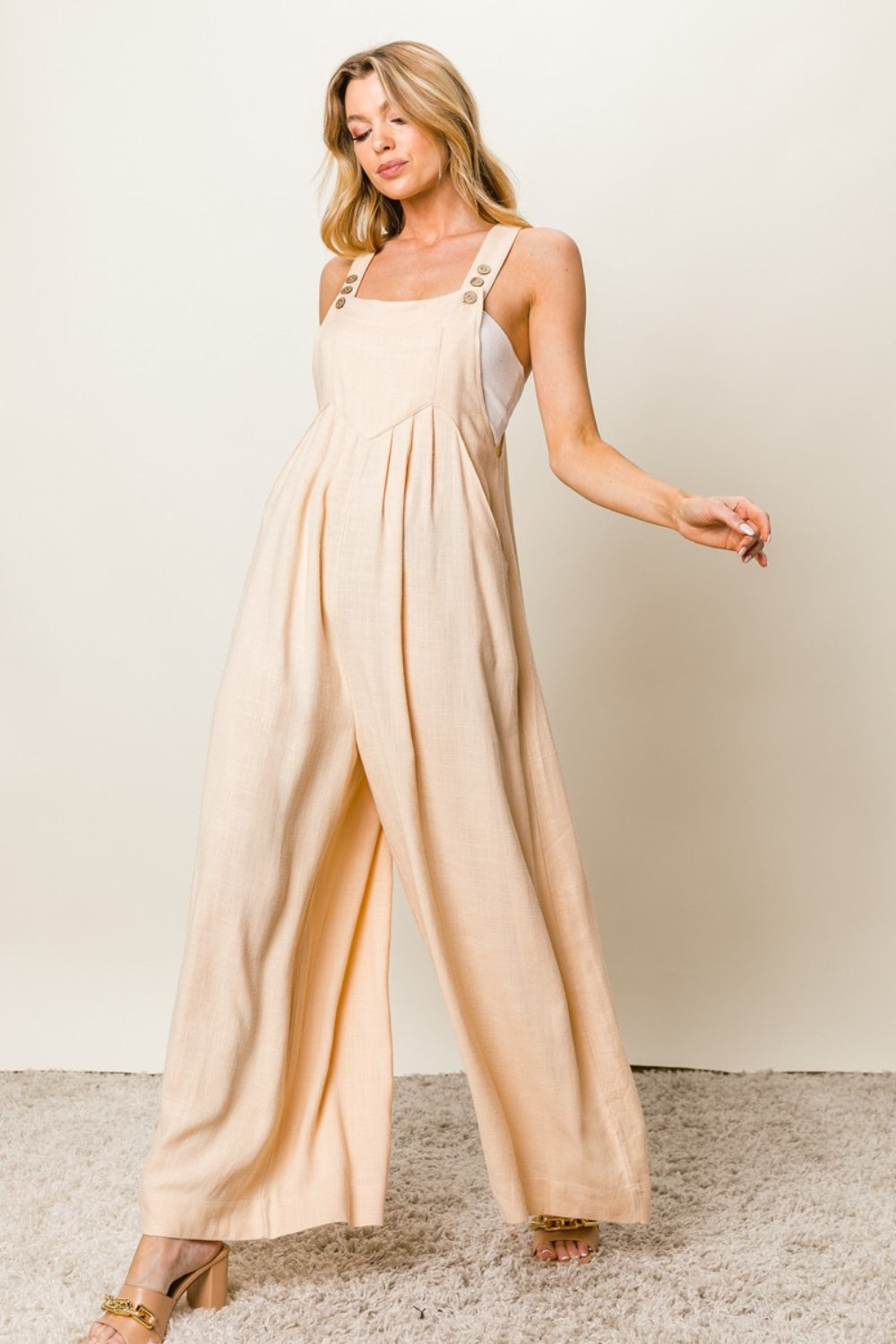 A person stands on a carpeted floor, wearing the BiBi Texture Sleeveless Wide Leg Jumpsuit in cream and beige high-heeled shoes.