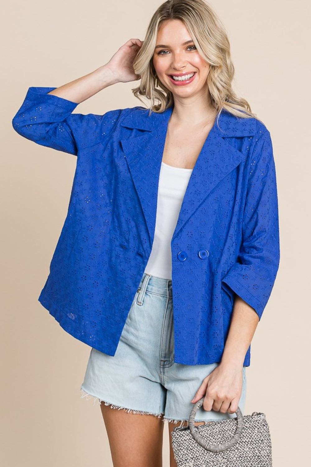 Person smiling, wearing a Culture Code Double Breasted Eyelet Jacket with Pockets over a white top and denim shorts, holding a small textured handbag.