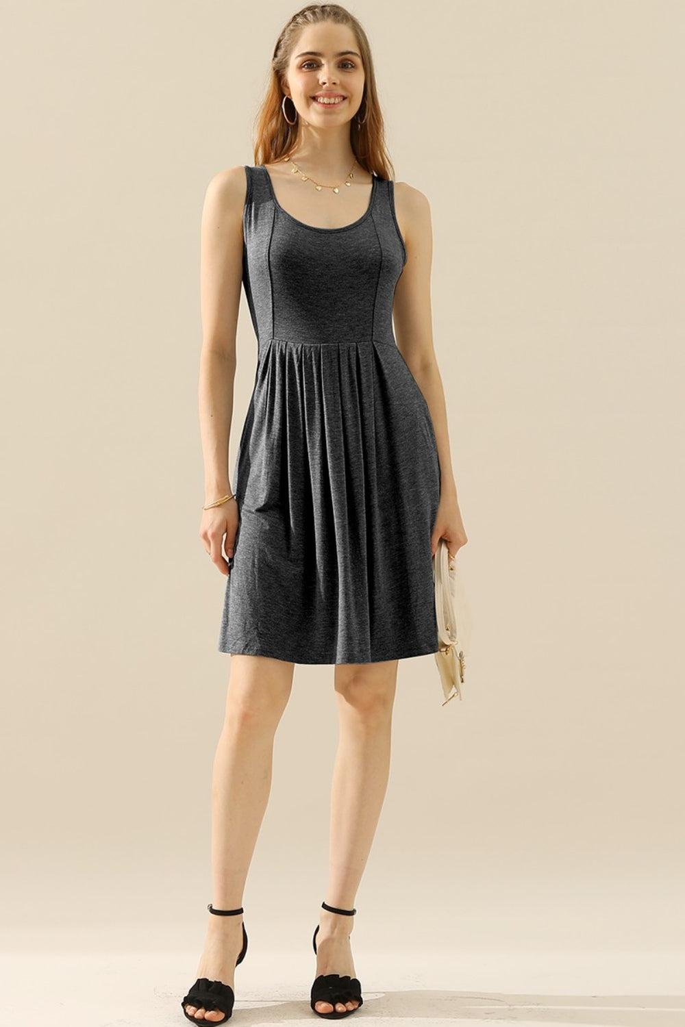 A woman in the Doublju Full Size Round Neck Ruched Sleeveless Dress with Pockets, featuring a stretchy gray fabric, smiles while holding a quilted white purse. She is wearing a gold necklace and bracelet.