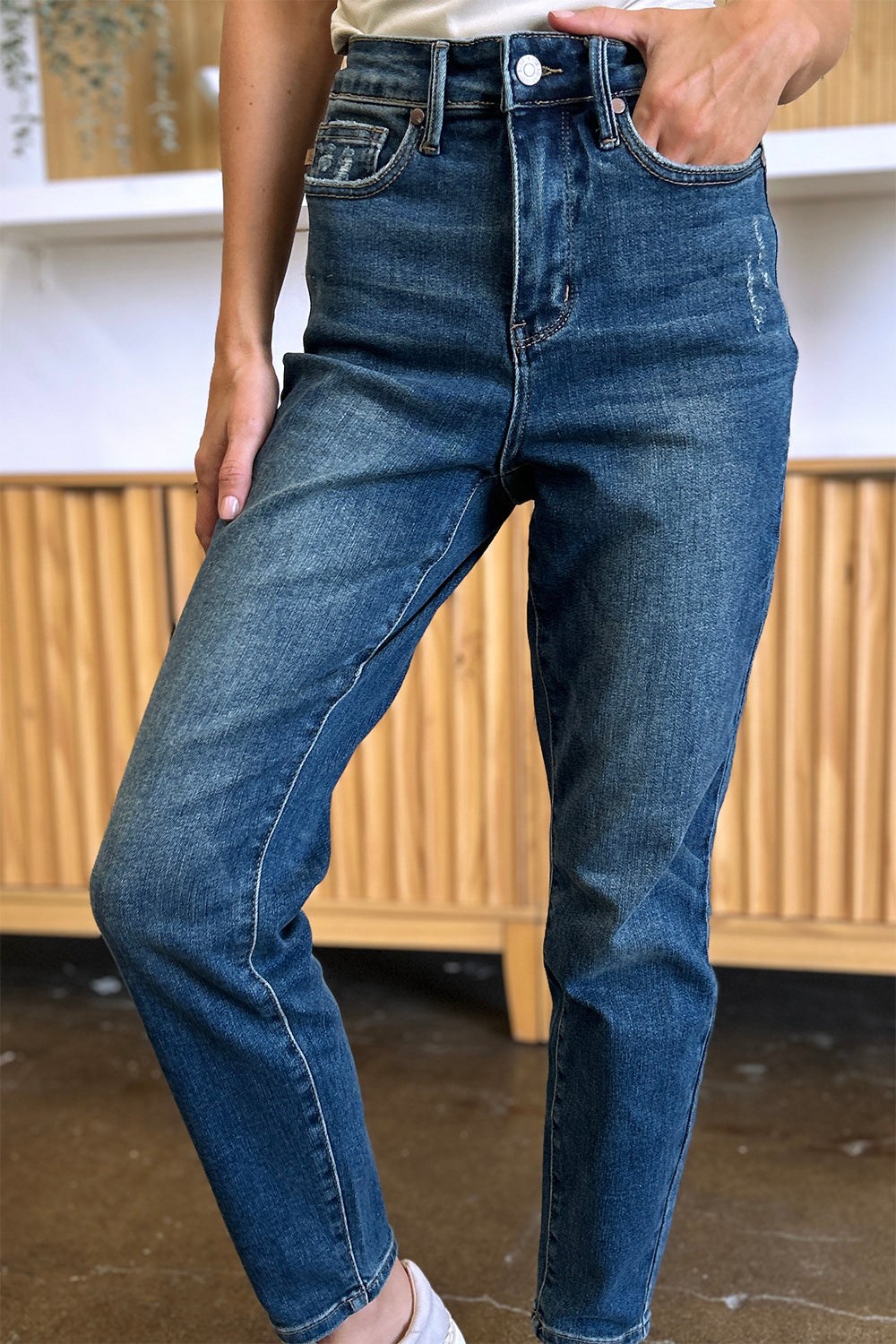 Person wearing Judy Blue Full Size Tummy Control High Waist Slim Jeans in blue and a white shirt, with their hands in their pockets. They are standing indoors on a polished concrete floor with a wooden cabinet and plant in the background.